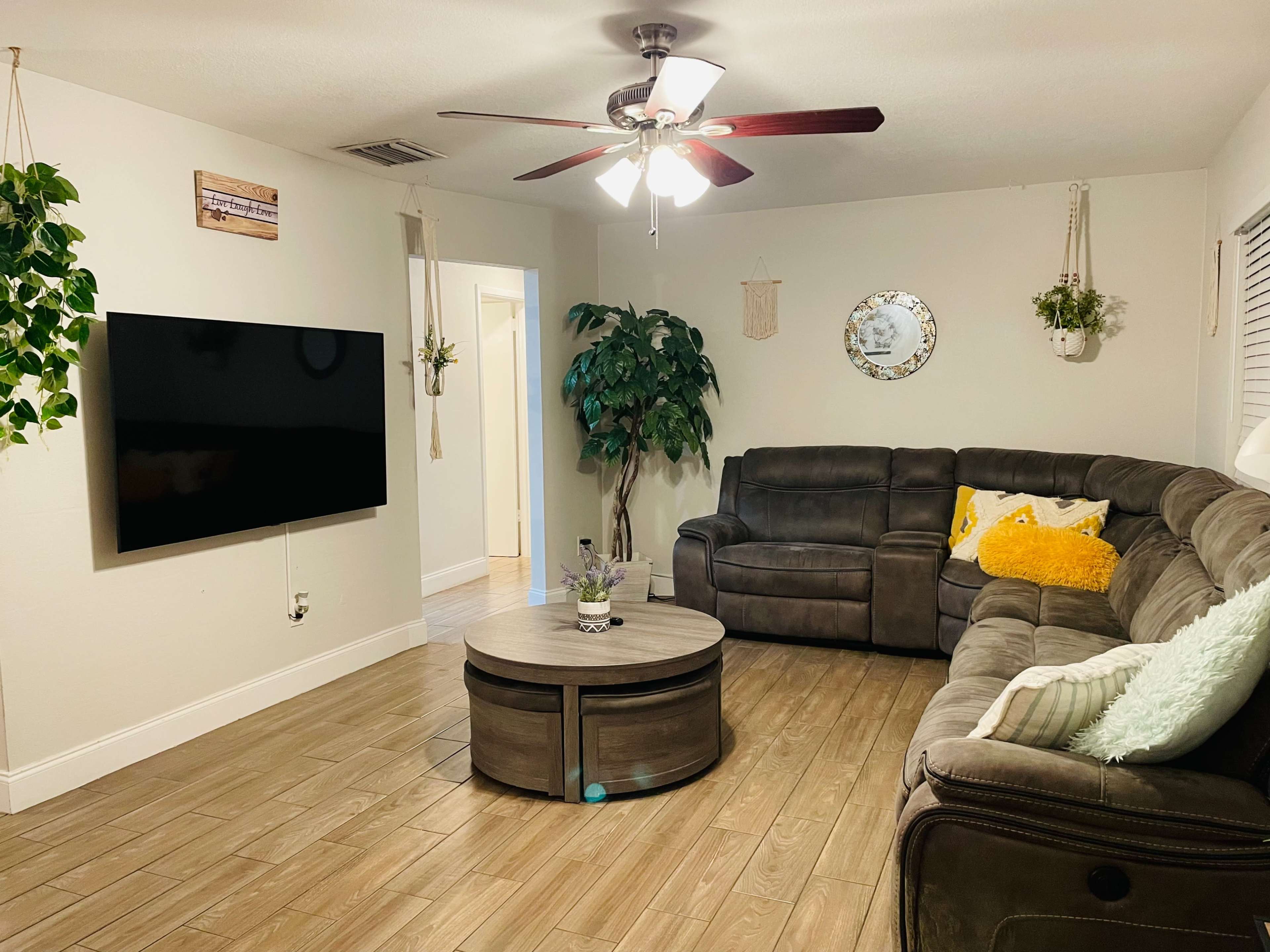 A living room with a gray sectional sofa, a round wooden coffee table, a mounted flat-screen TV, and decorative plants hanging from the walls.