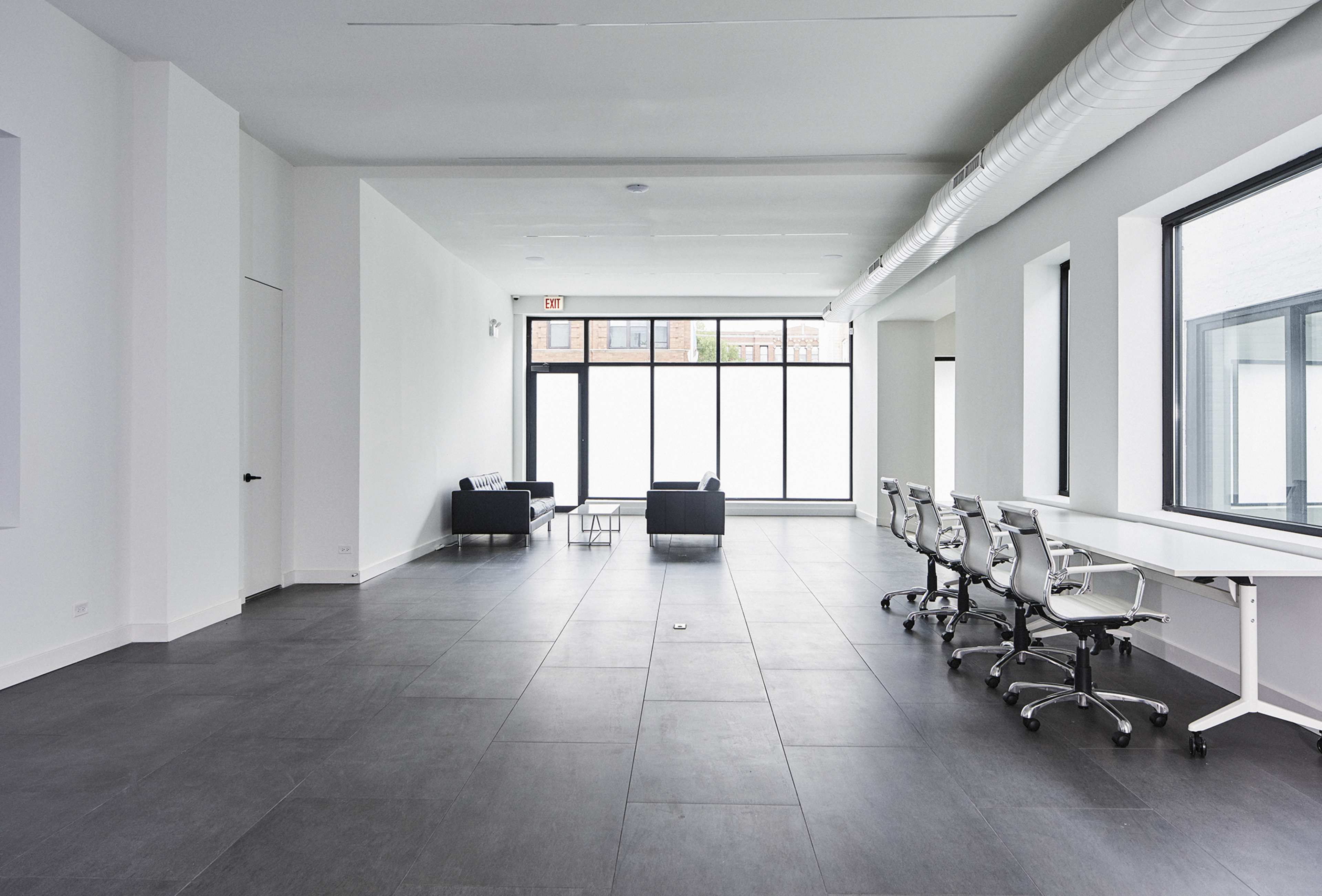 A modern, minimalist office space with two black couches and several white desks with rolling chairs, illuminated by natural light from large windows.