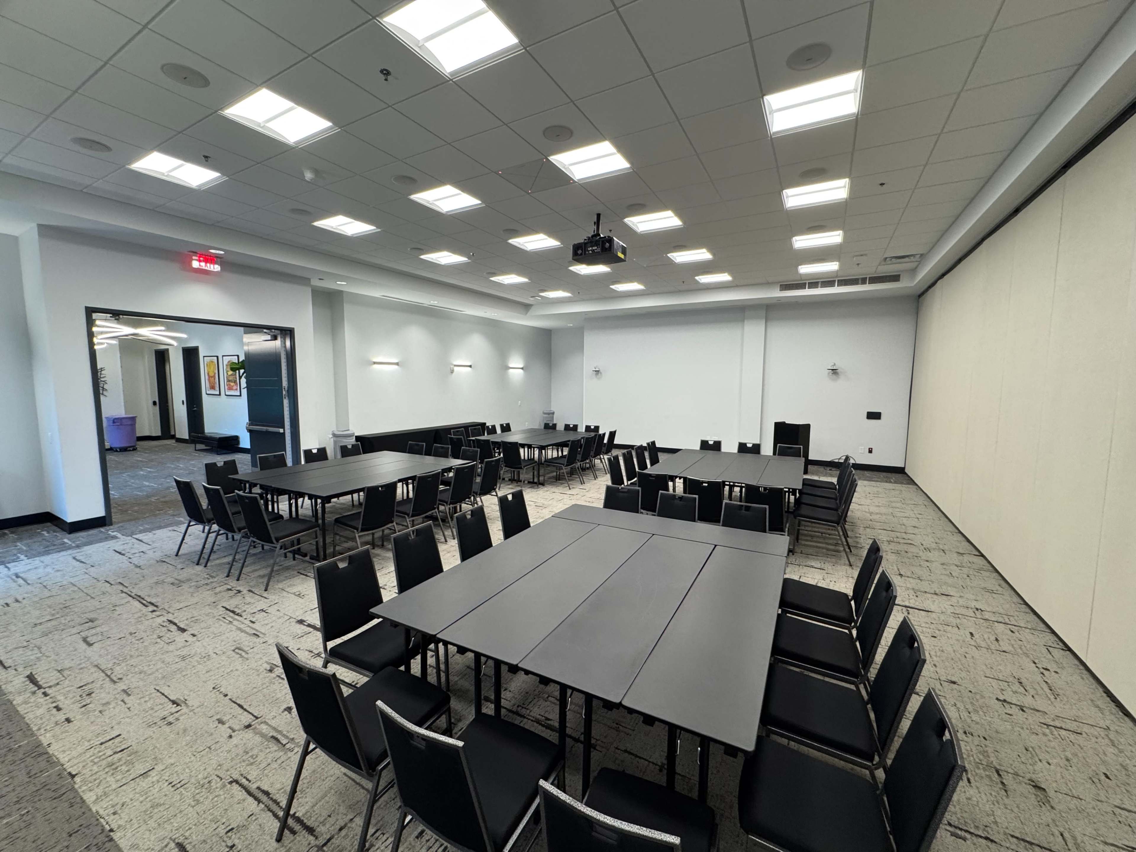 A spacious conference room features multiple rectangular tables arranged in a U-shape, surrounded by black chairs, with bright overhead lighting.