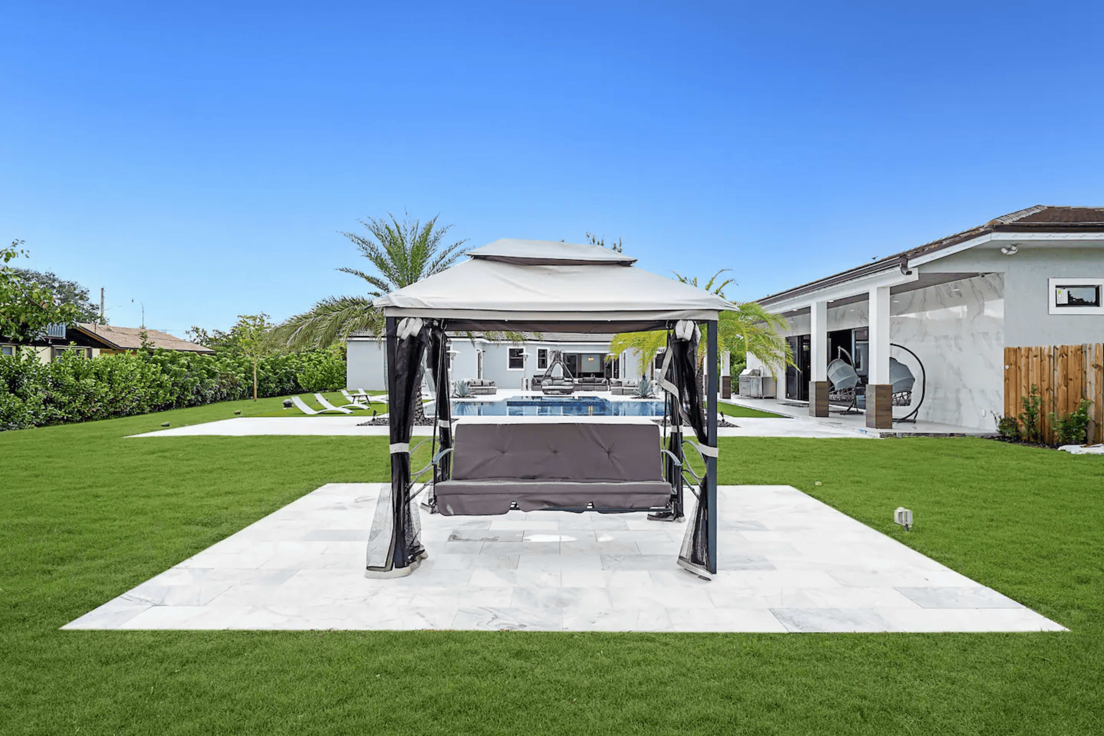 A gazebo with curtains is situated on a marble patio, surrounded by green grass and palm trees, with a swimming pool and house visible in the background.