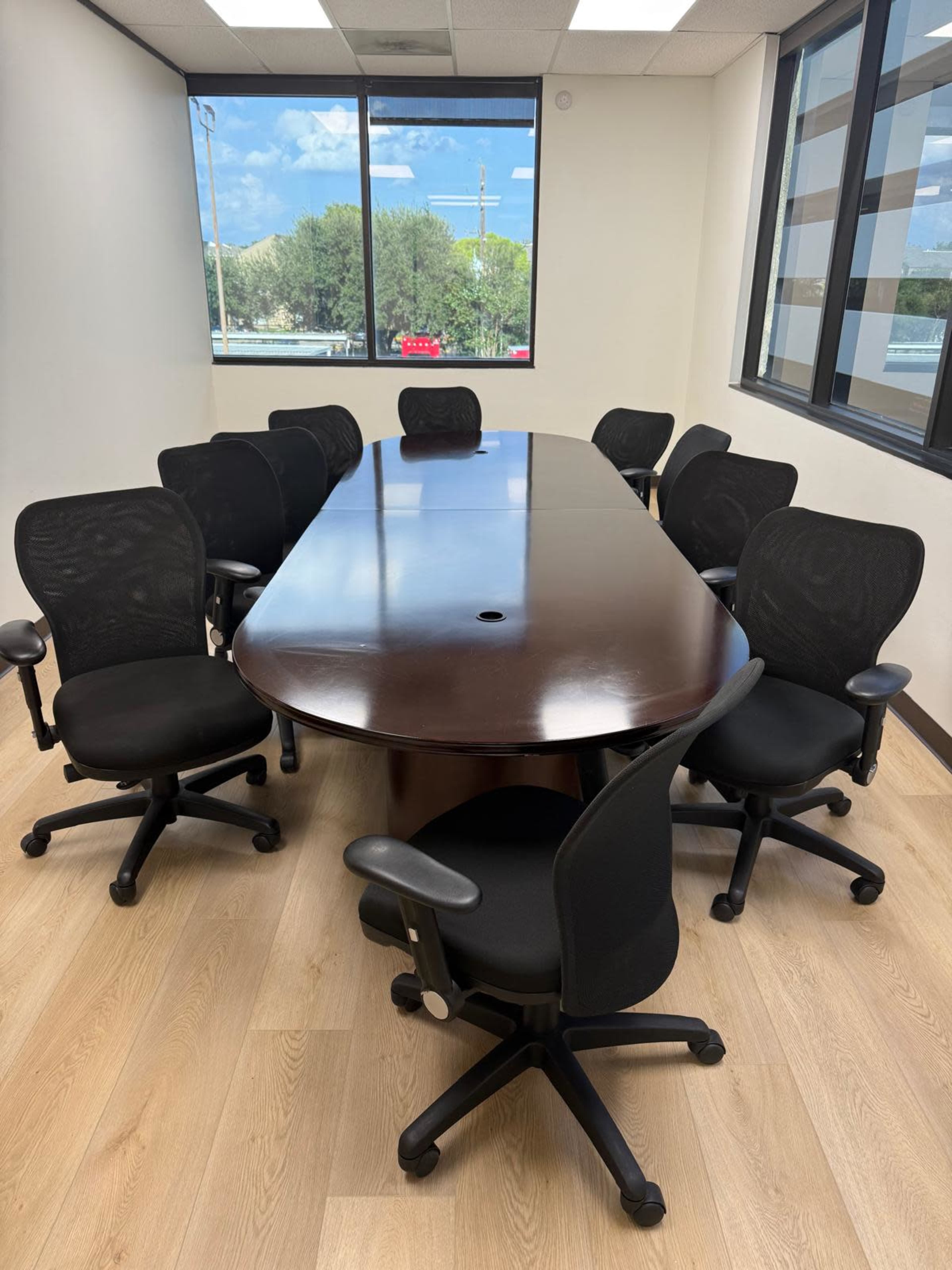 A large, oval conference table surrounded by eight black office chairs in a well-lit room with windows.