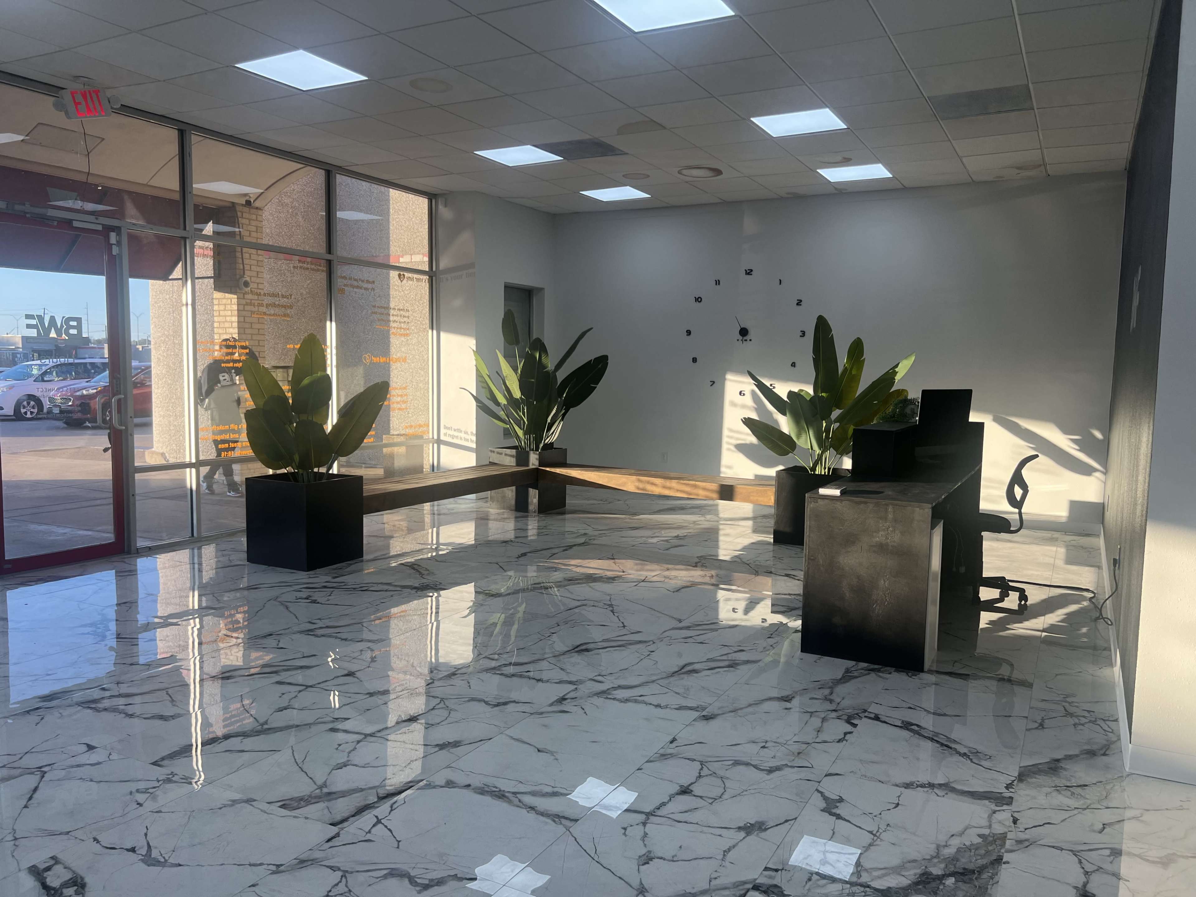 The image shows a bright, modern lobby with marble flooring, two large potted plants, and a reception desk against a wall of windows.