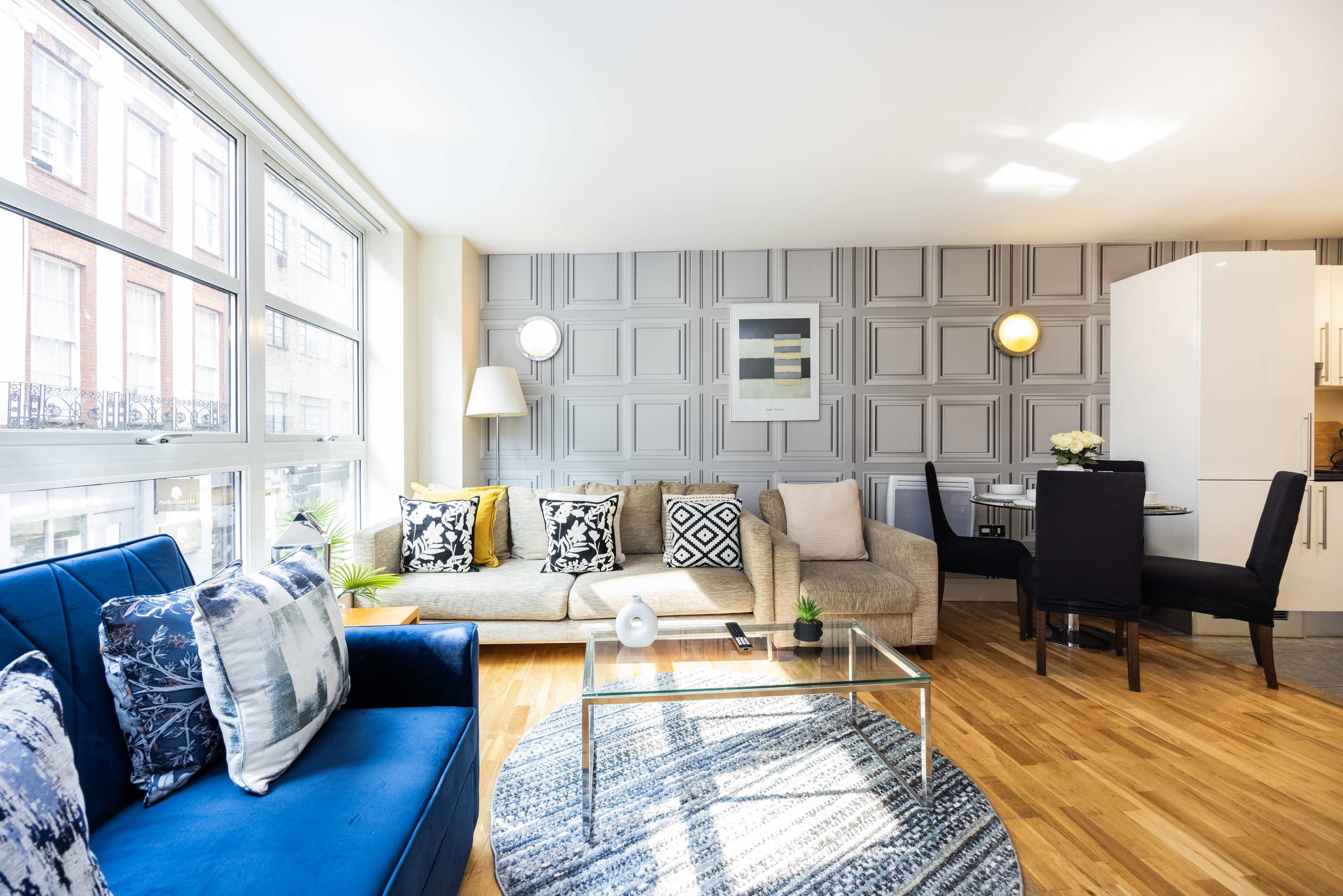 A modern living room features a sofa, blue chairs, a glass coffee table, and a dining area with a backdrop of patterned walls and large windows allowing natural light.