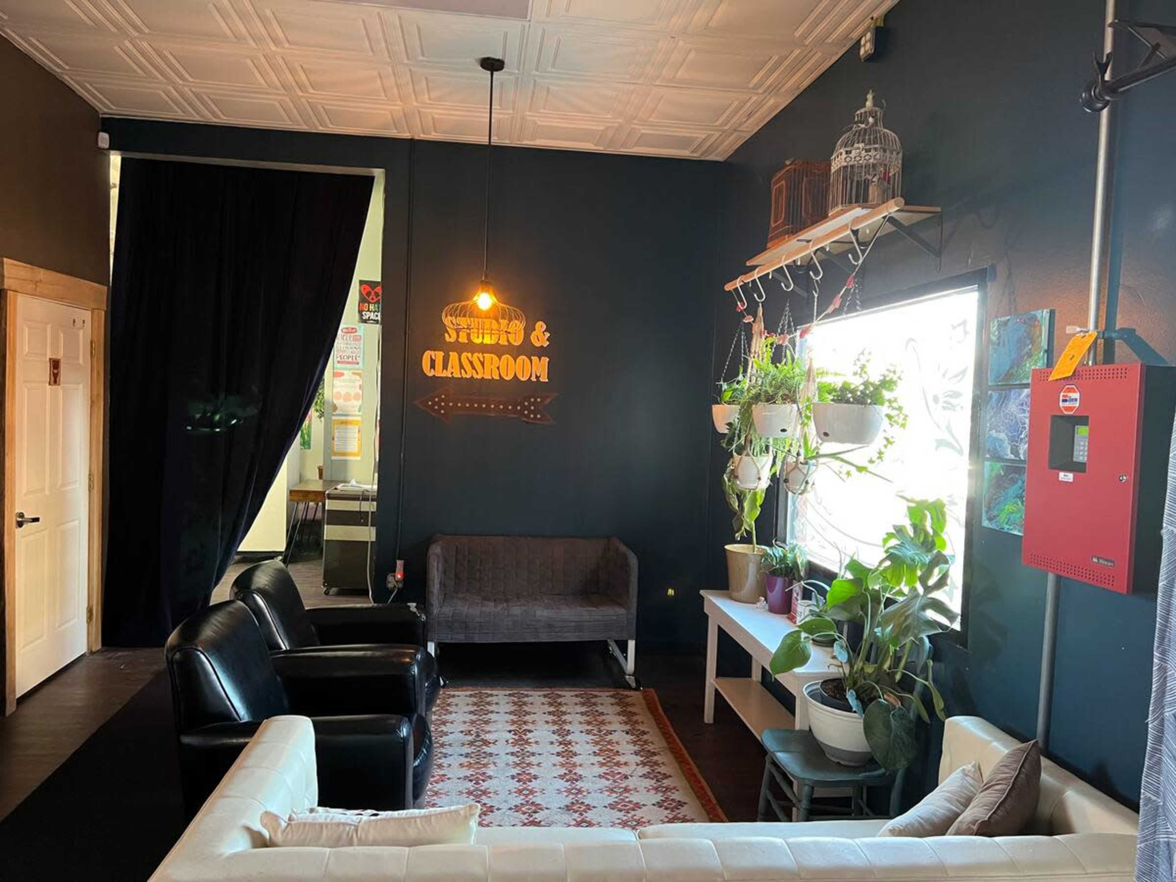 A cozy interior space with black chairs and a white couch, featuring plants by the window and a sign indicating "STUDIO & CLASSROOM."