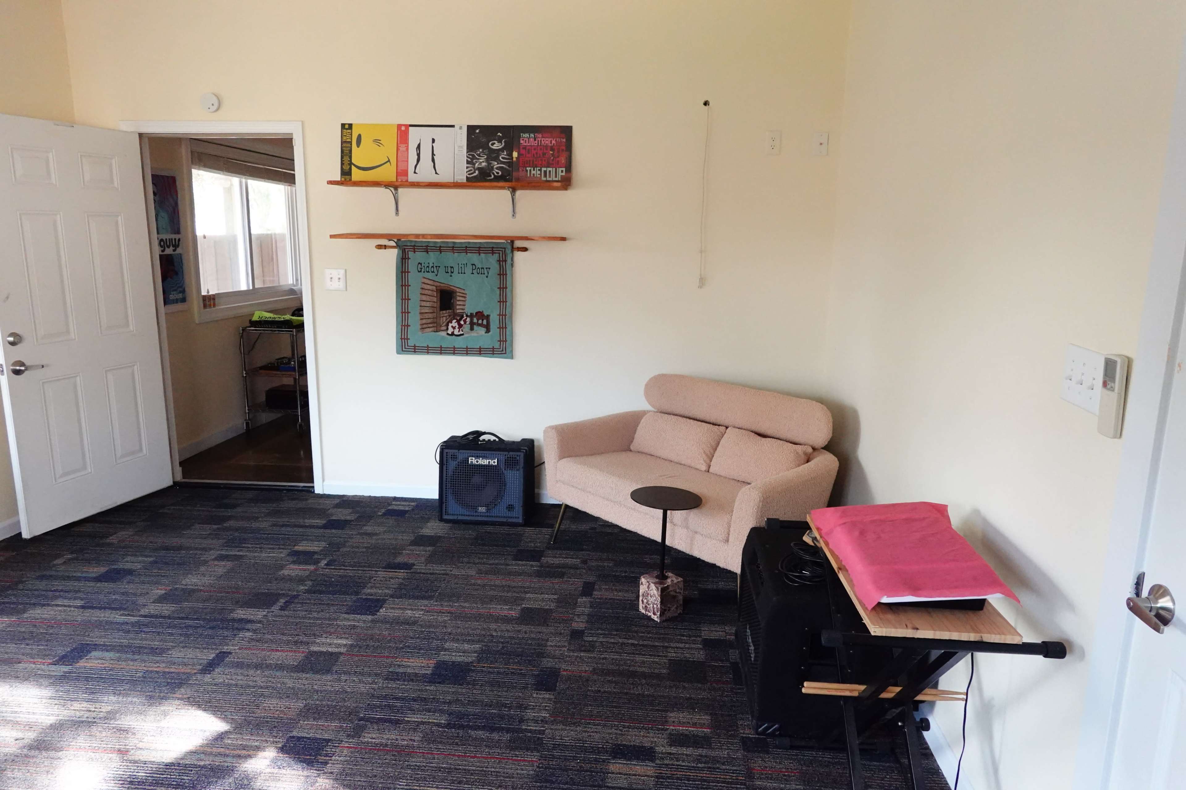 A small room with a beige couch, a side table, an amplifier on the floor, and several colorful art pieces on the walls.