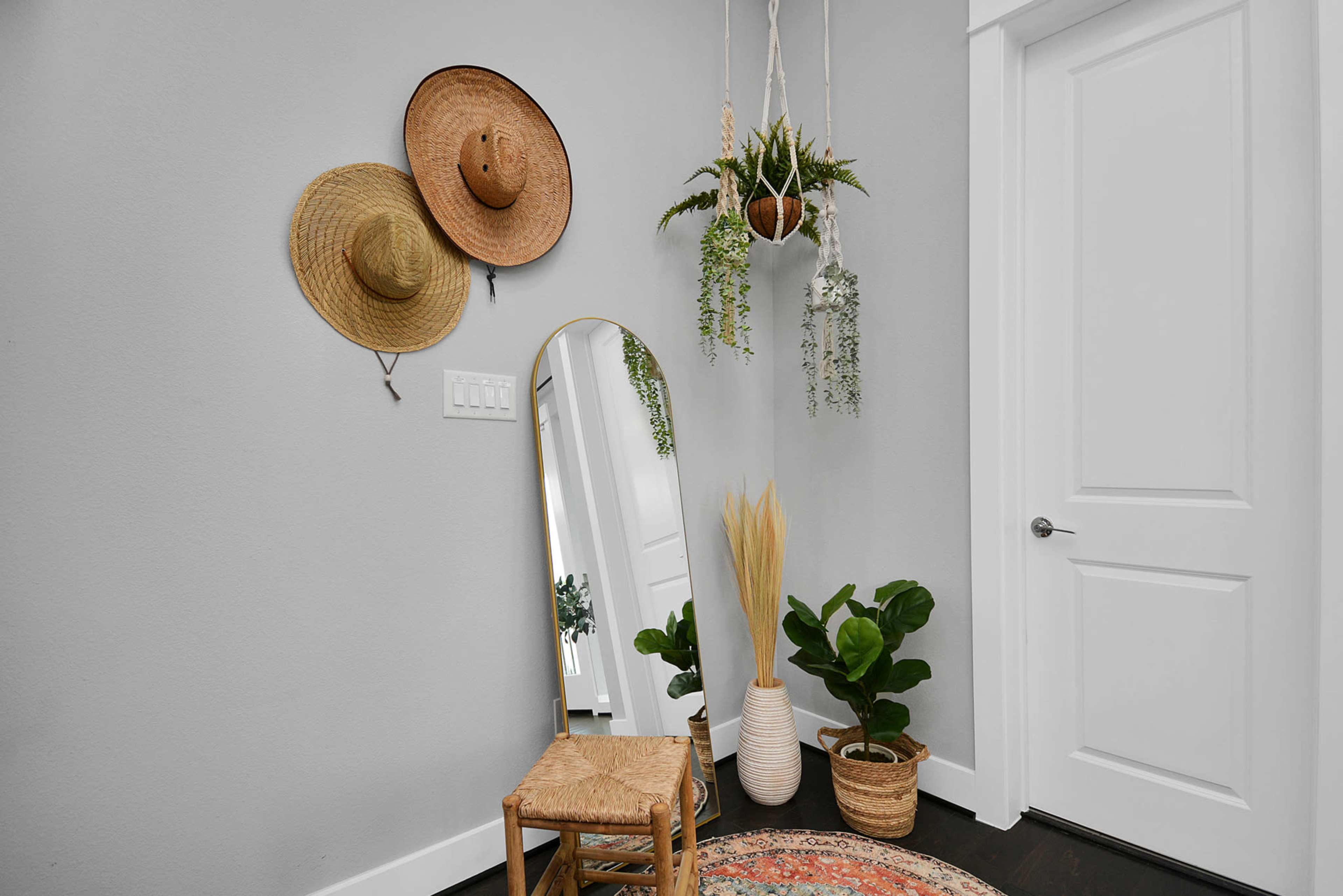 A hallway features two straw hats hanging on the wall beside a mirror, a chair, and potted plants, with a door visible in the background.