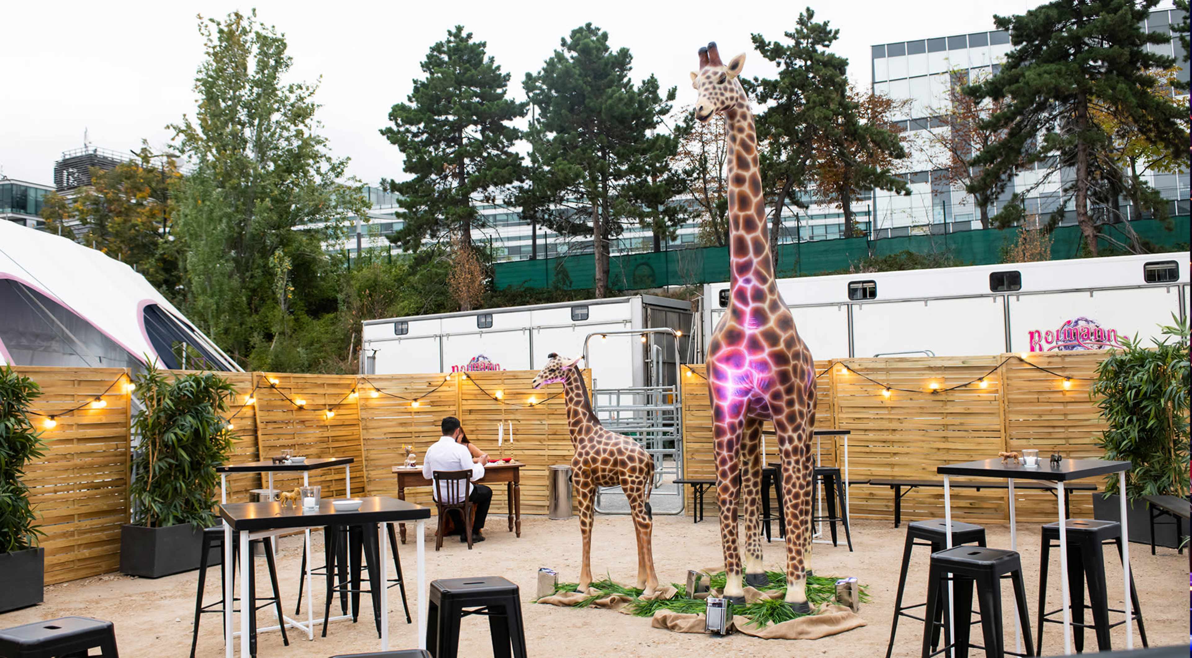 The scene features a fenced outdoor space with two large giraffe statues, surrounded by tables and chairs, and a person sitting nearby.