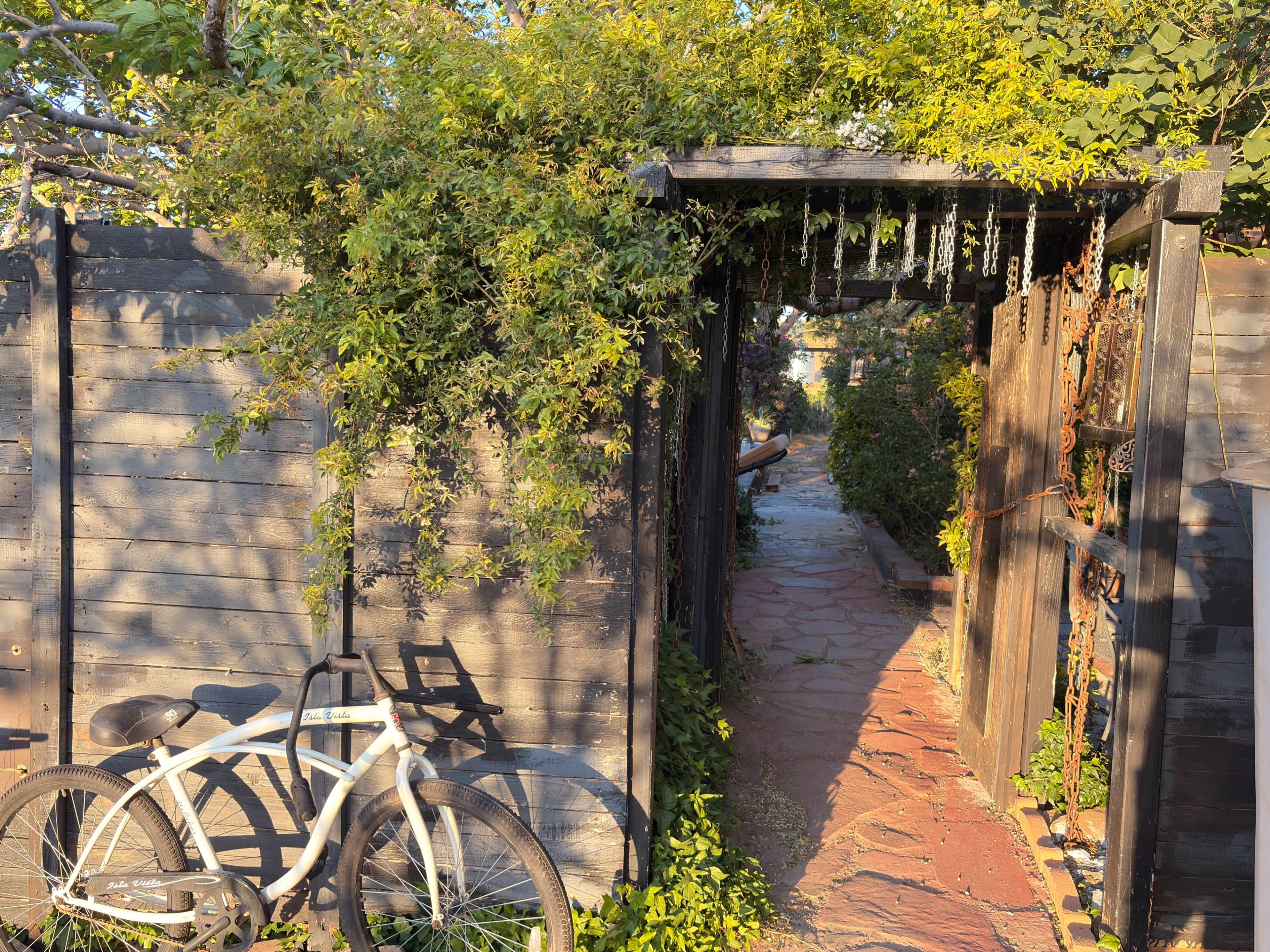 A bike leans against a fence covered in greenery beside an open wooden gate leading to a stone path.