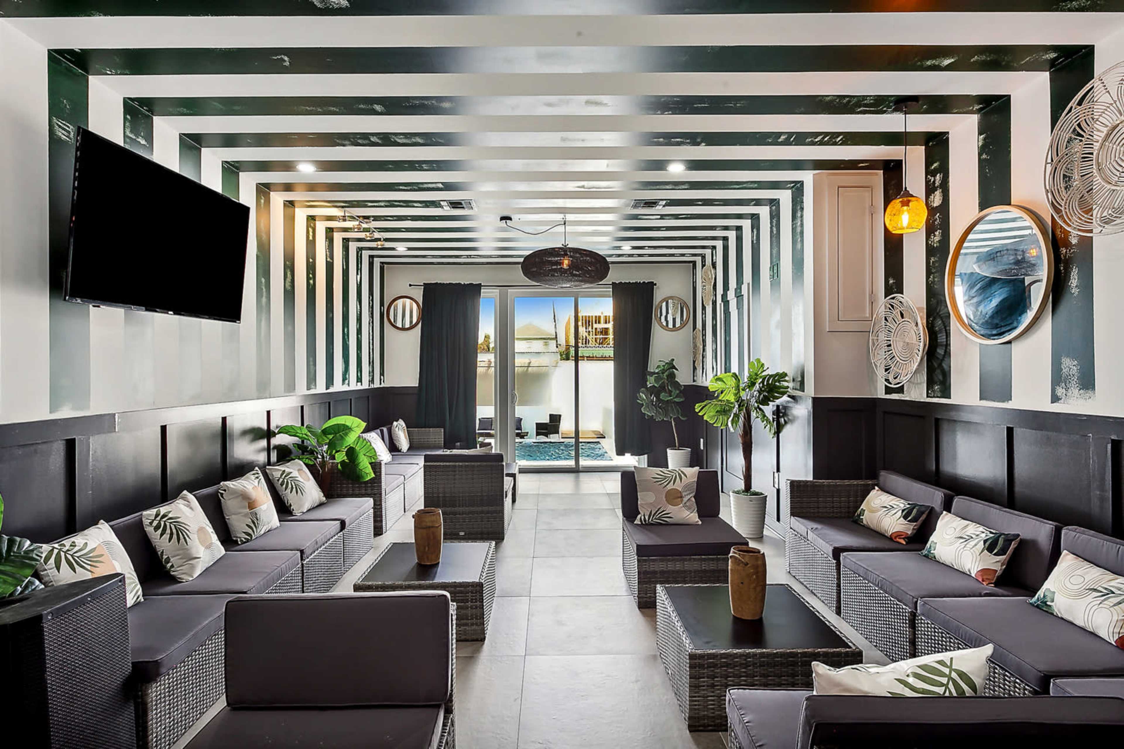 A modern lounge area with dark furniture, potted plants, and large windows overlooking a swimming pool.