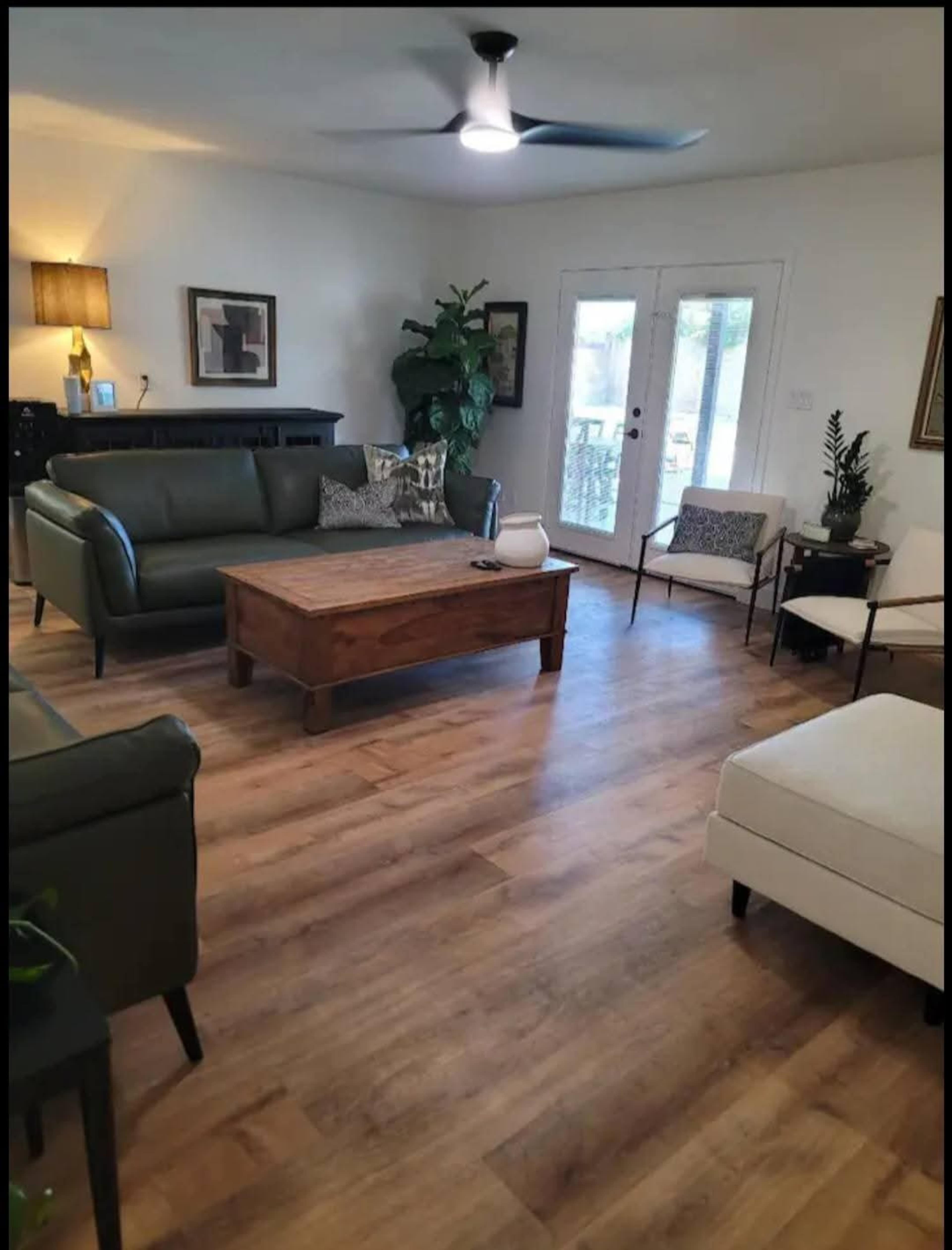 The image shows a living room featuring a dark green sofa, a wooden coffee table, and two armchairs near a set of double doors leading to an outdoor area.