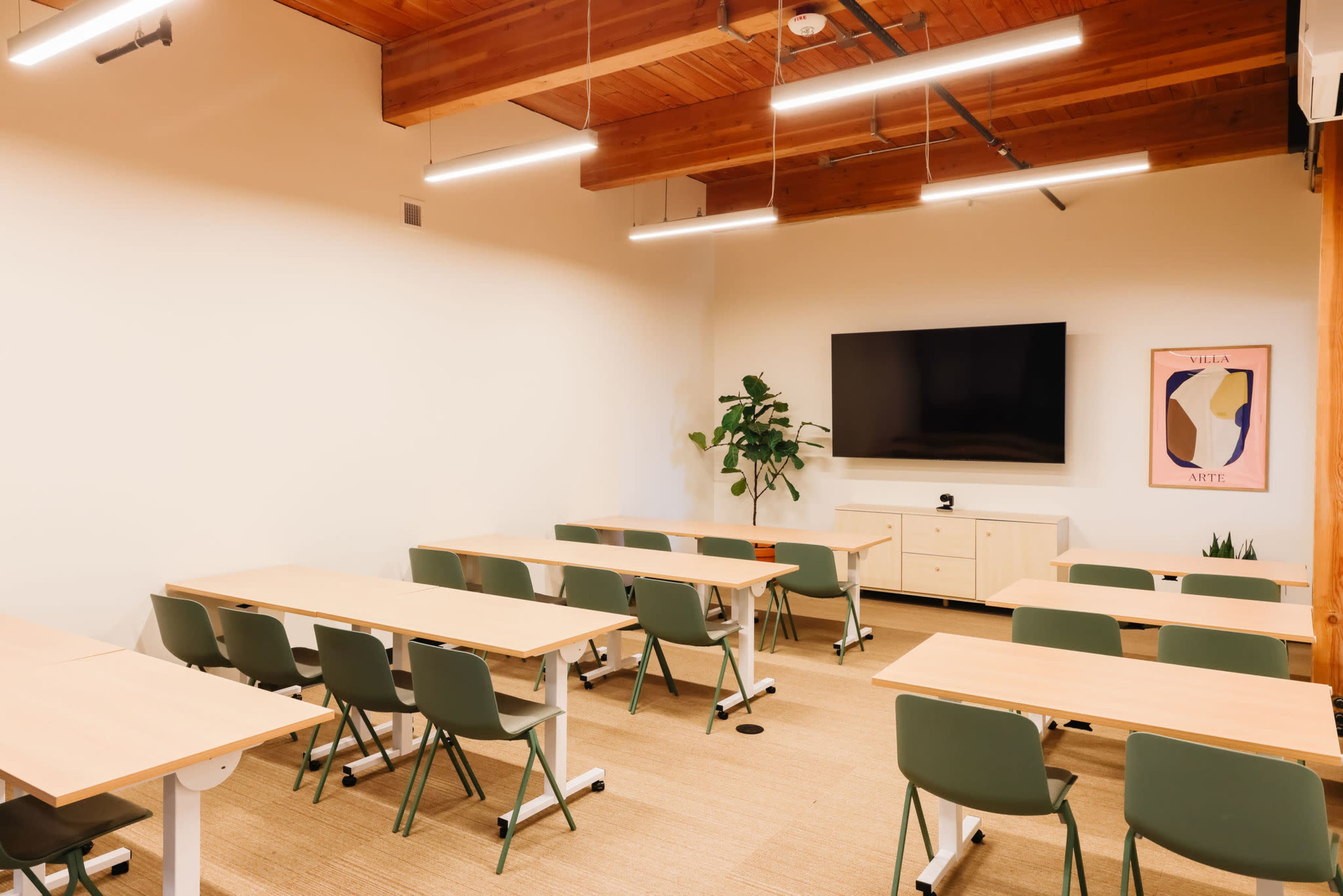 A modern classroom featuring several rows of tables and chairs, a large television mounted on the wall, and a green plant in one corner.