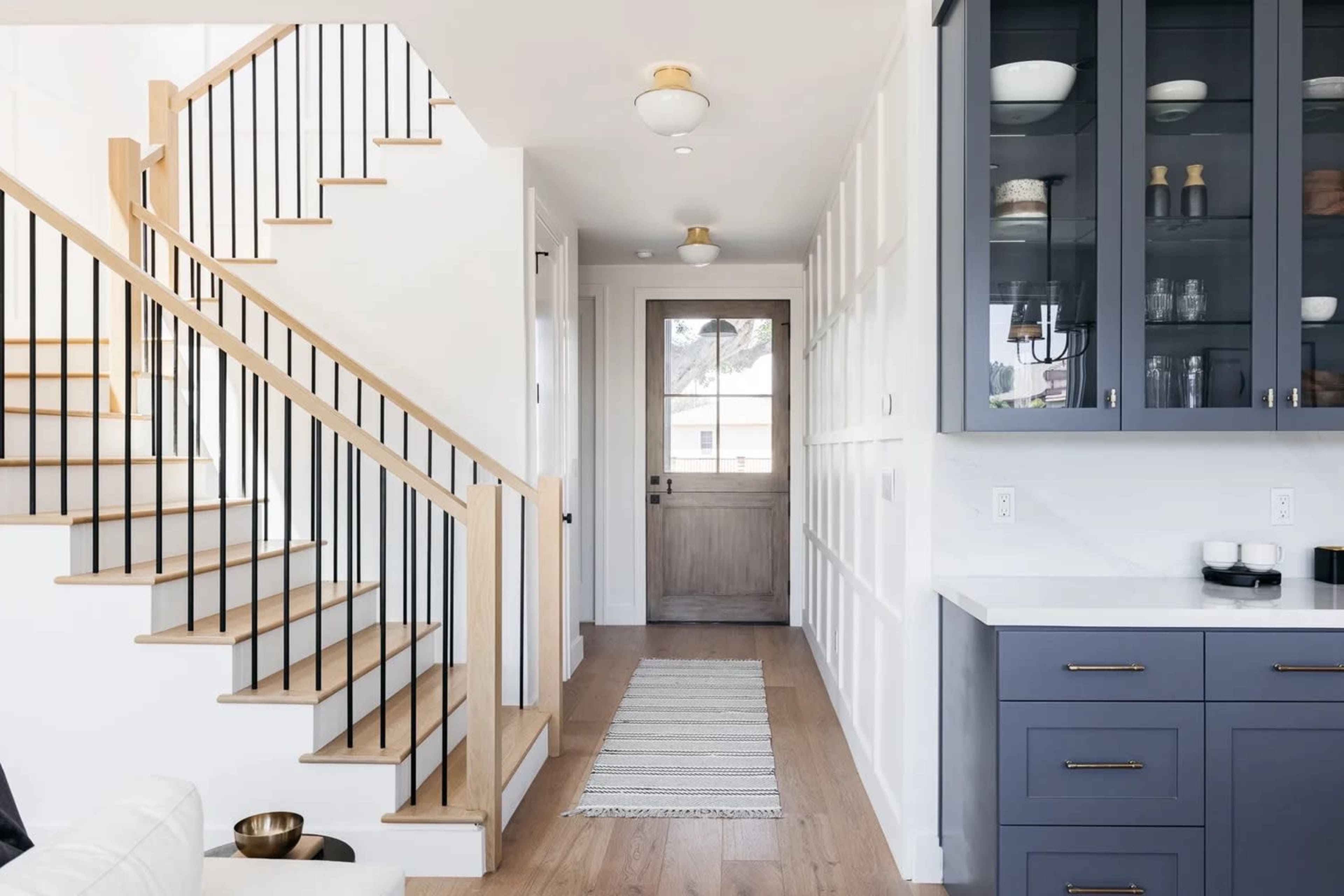 A bright entryway features a staircase with wooden steps and black railings, leading to a door, alongside a wall of blue cabinetry and a narrow white runner on the floor.