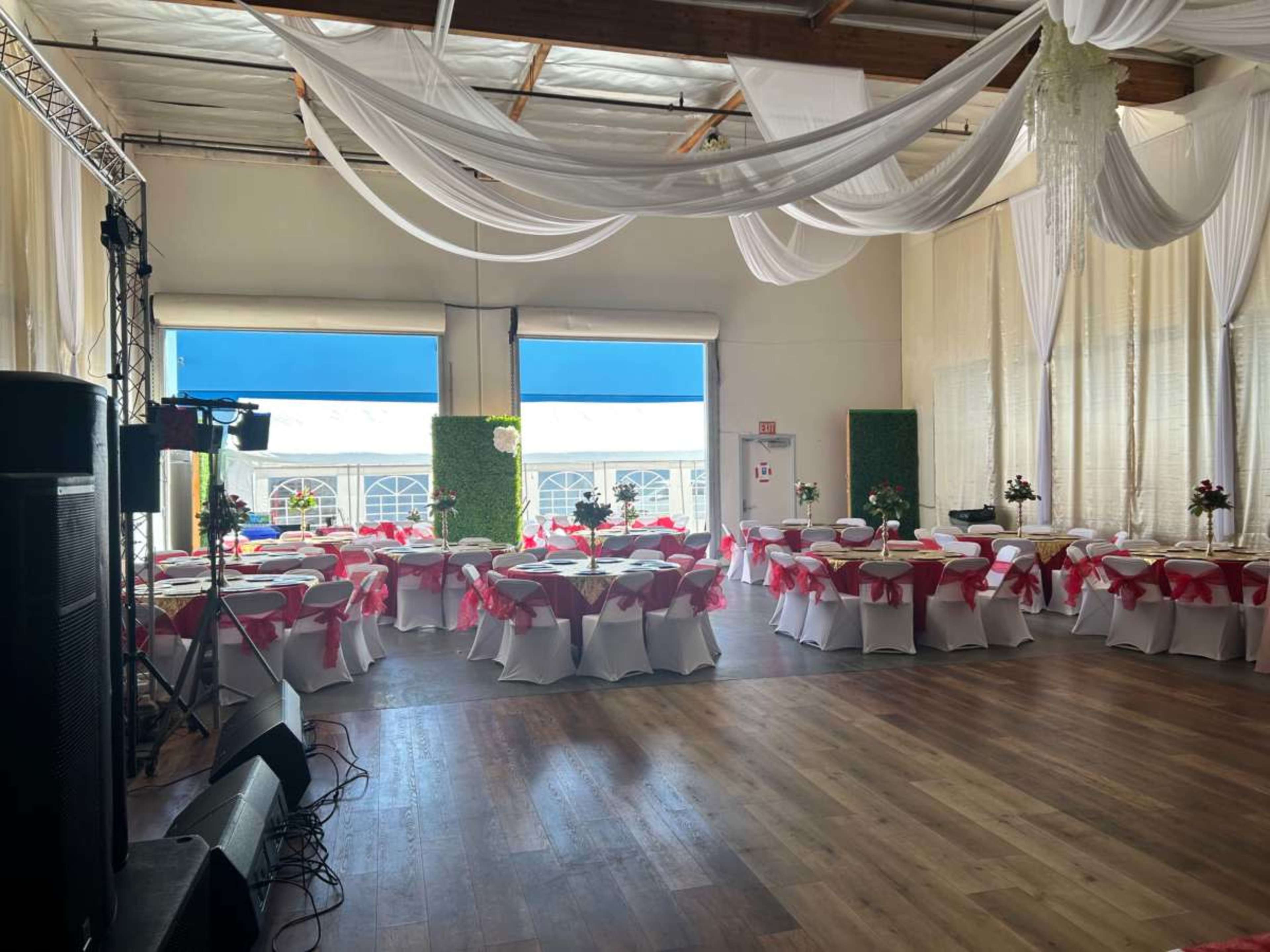 The image shows a spacious indoor event venue decorated with tables covered in white tablecloths and pink ribbons, with floral centerpieces and large windows letting in natural light.