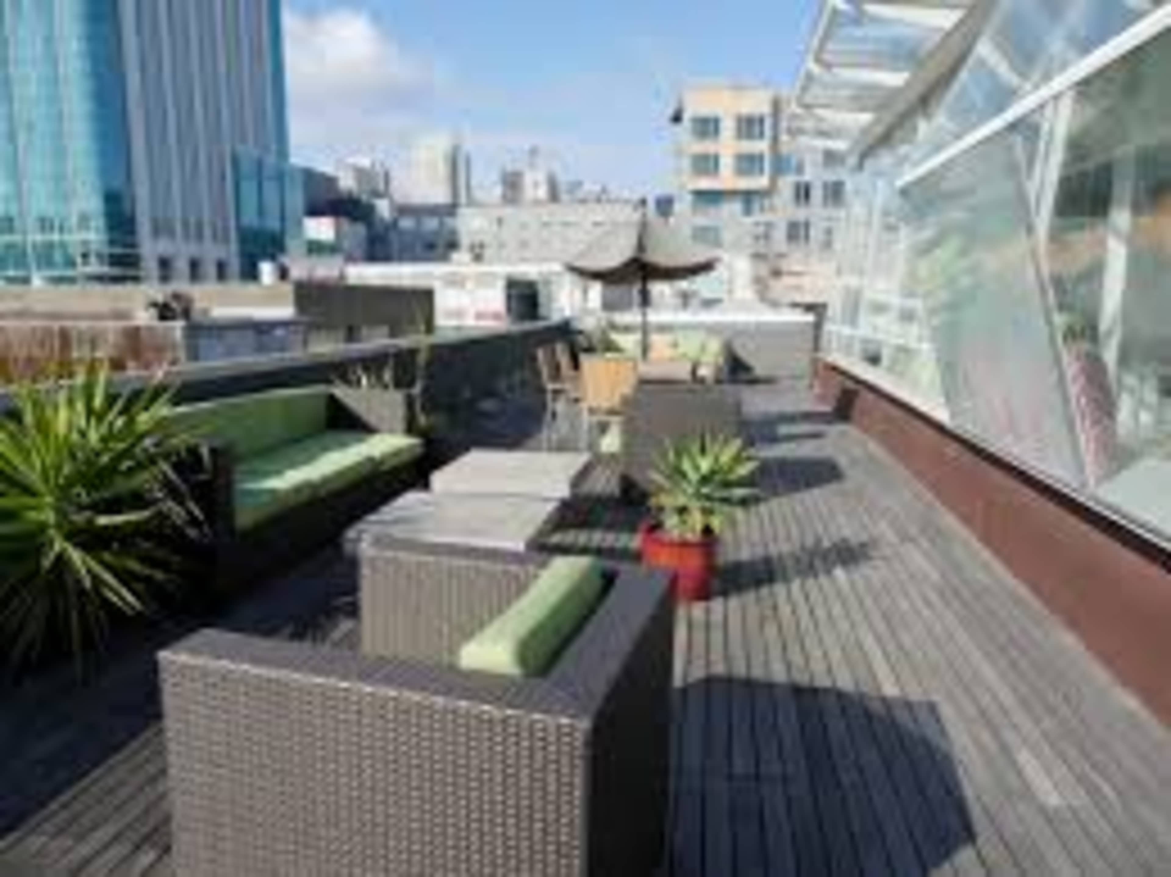 A modern rooftop terrace with seating, small plants, and a city skyline in the background.