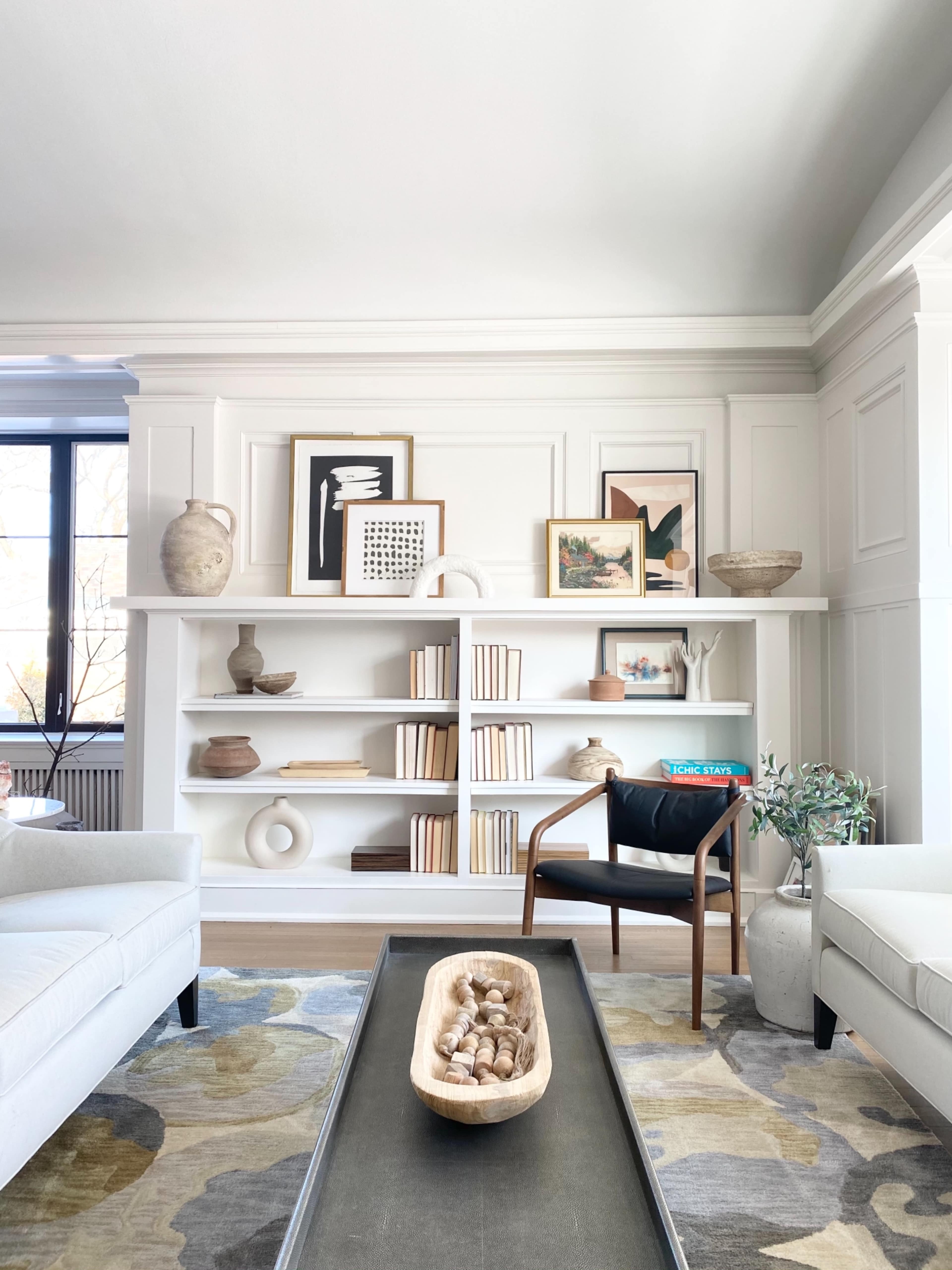 A styled living room features a white bookshelf with art and decorative objects, a wooden bowl on a coffee table, and a modern chair.