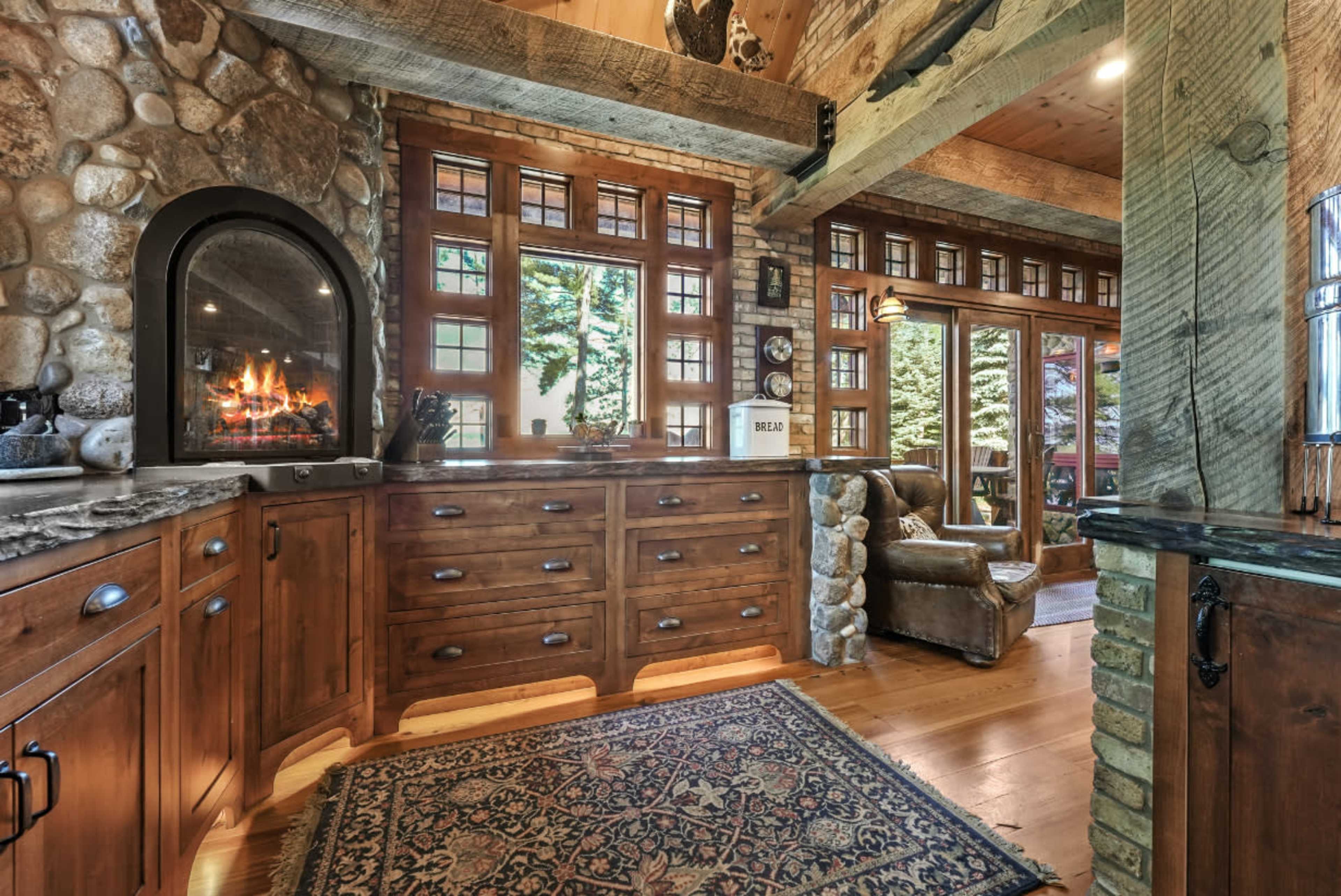 The image shows a rustic interior of a cabin featuring wooden cabinetry, a stone fireplace, and large windows that offer a view of the outdoors.