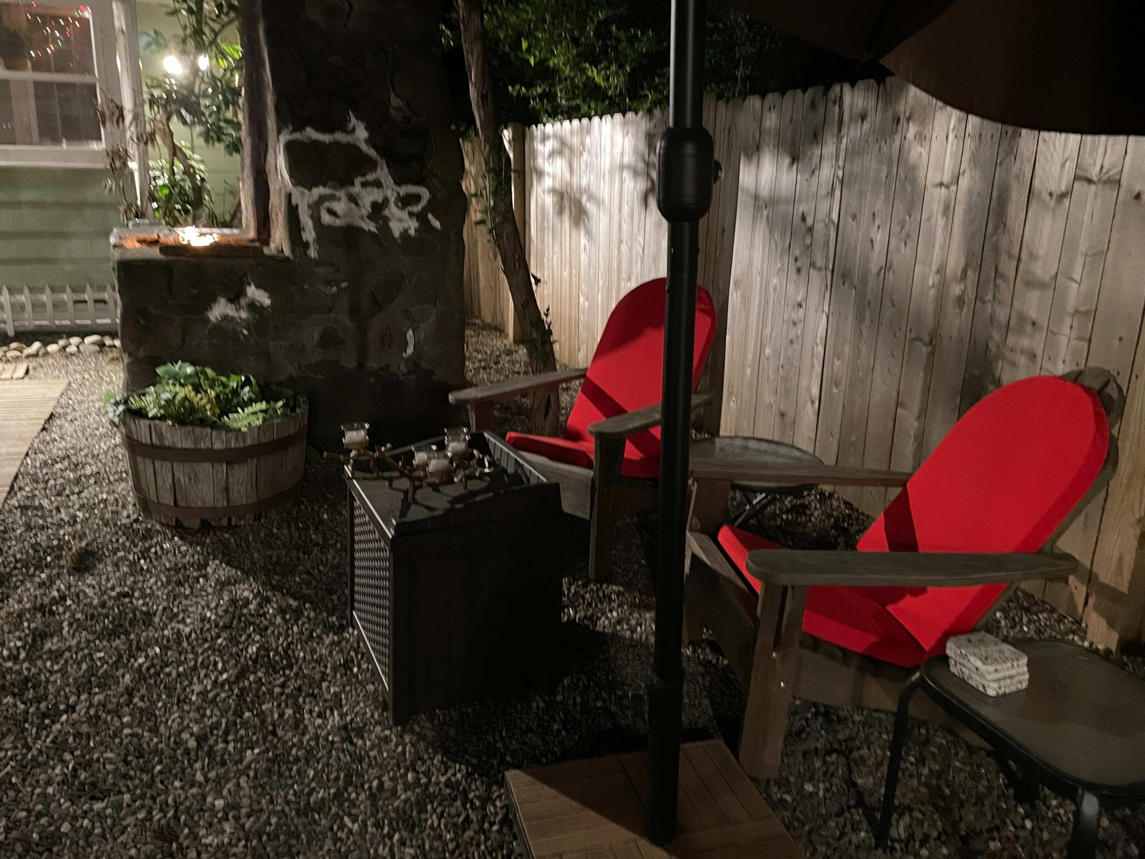 The image shows a cozy outdoor seating area with two red chairs next to a stone wall, a small table, and dim lighting.