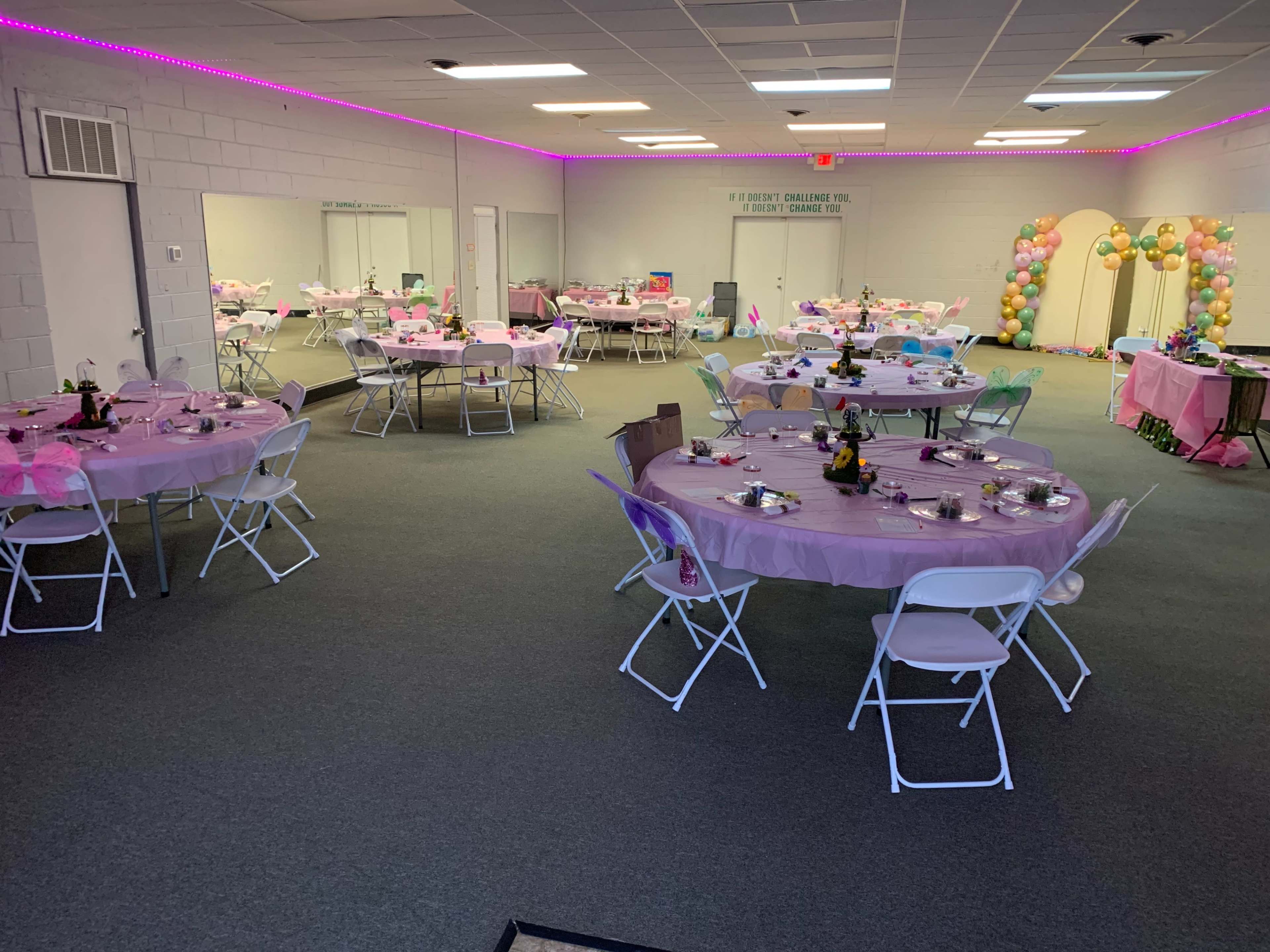 The image shows an event space arranged with multiple tables covered in pink tablecloths, decorated with centerpieces, and colorful balloons arching at one end.