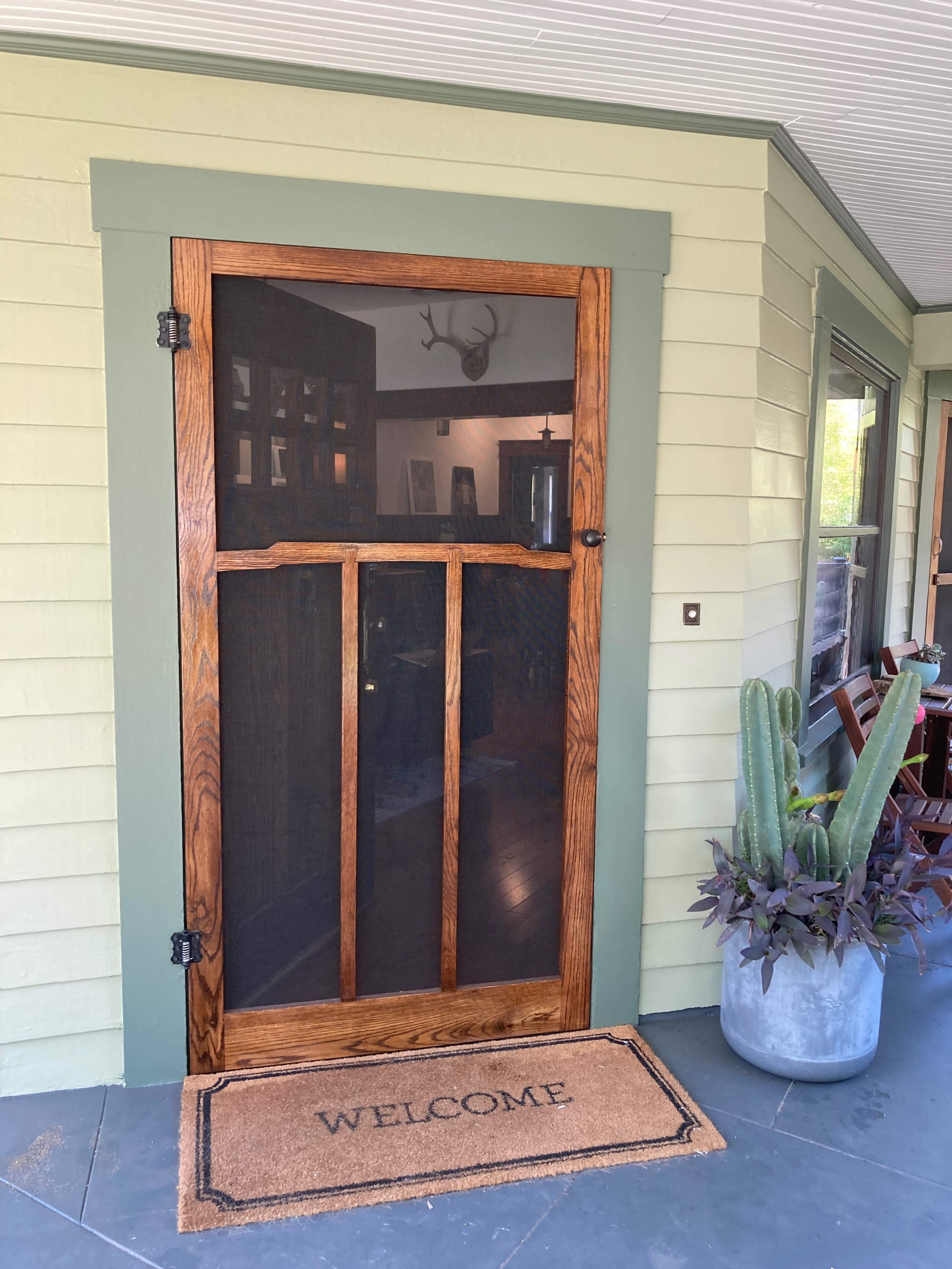 A wooden screen door with a "WELCOME" mat in front, set against a light green house exterior and a potted plant nearby.