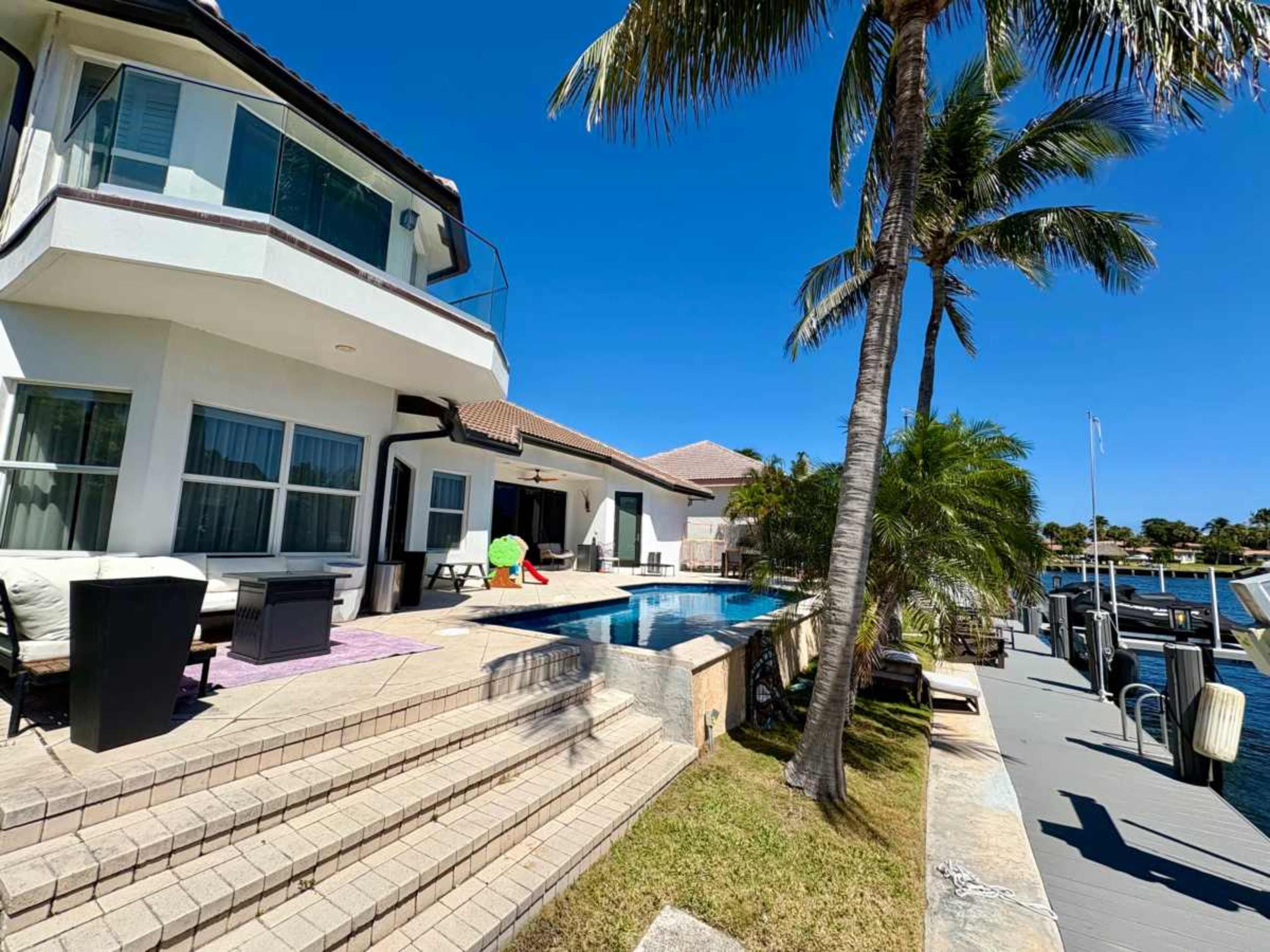 A modern house with large windows overlooks a swimming pool and is surrounded by palm trees and a waterfront.