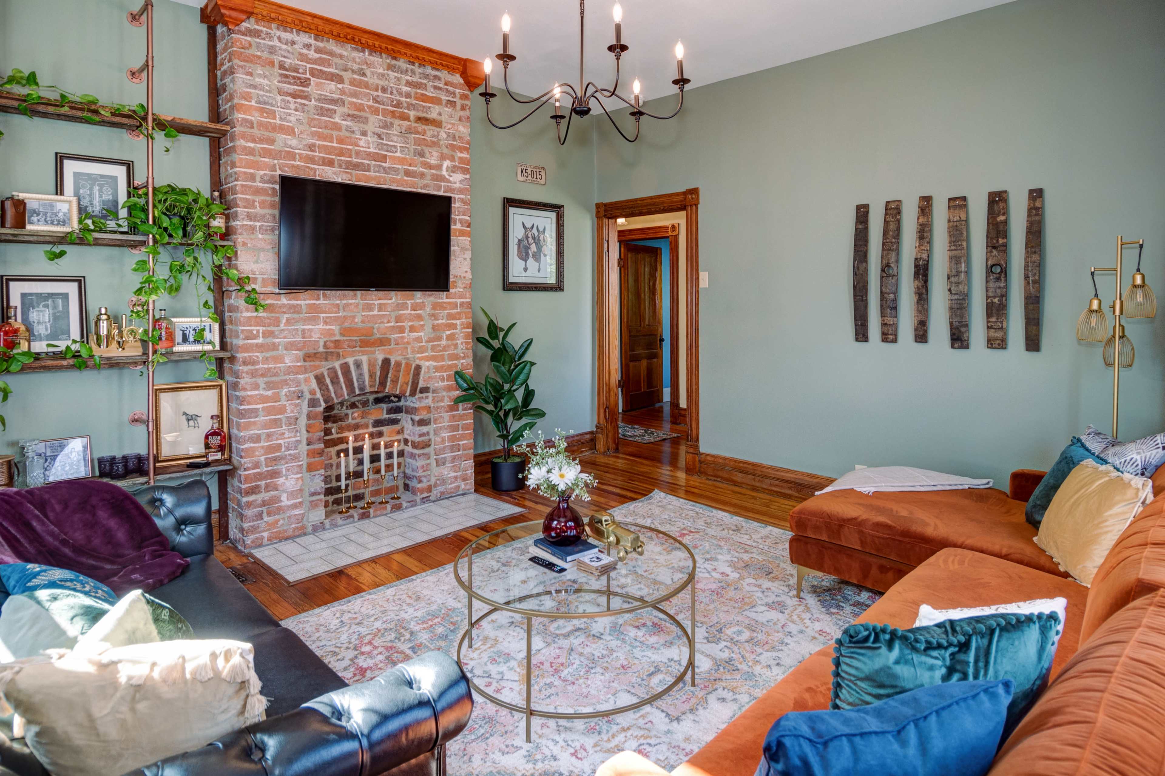 A living room with a brick fireplace, a mounted TV, and a mix of seating arrangements, including an orange sofa and a black leather couch.