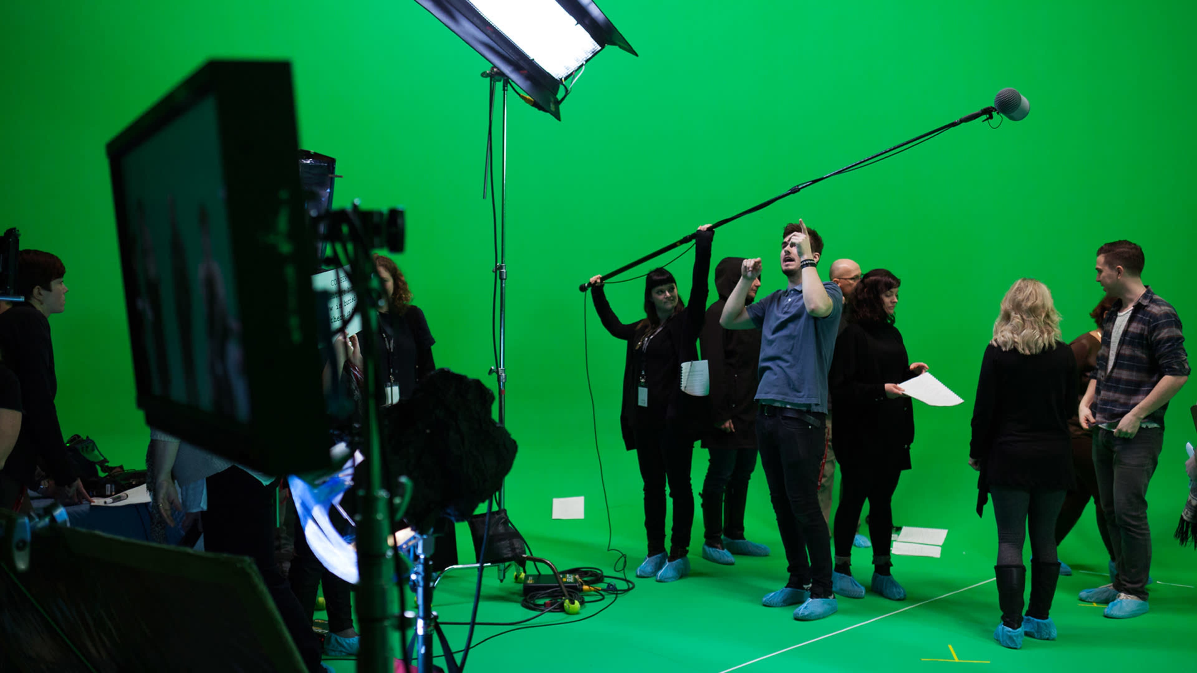 A group of people is gathered in a studio with a green screen, using lighting equipment and a microphone while reviewing a script.
