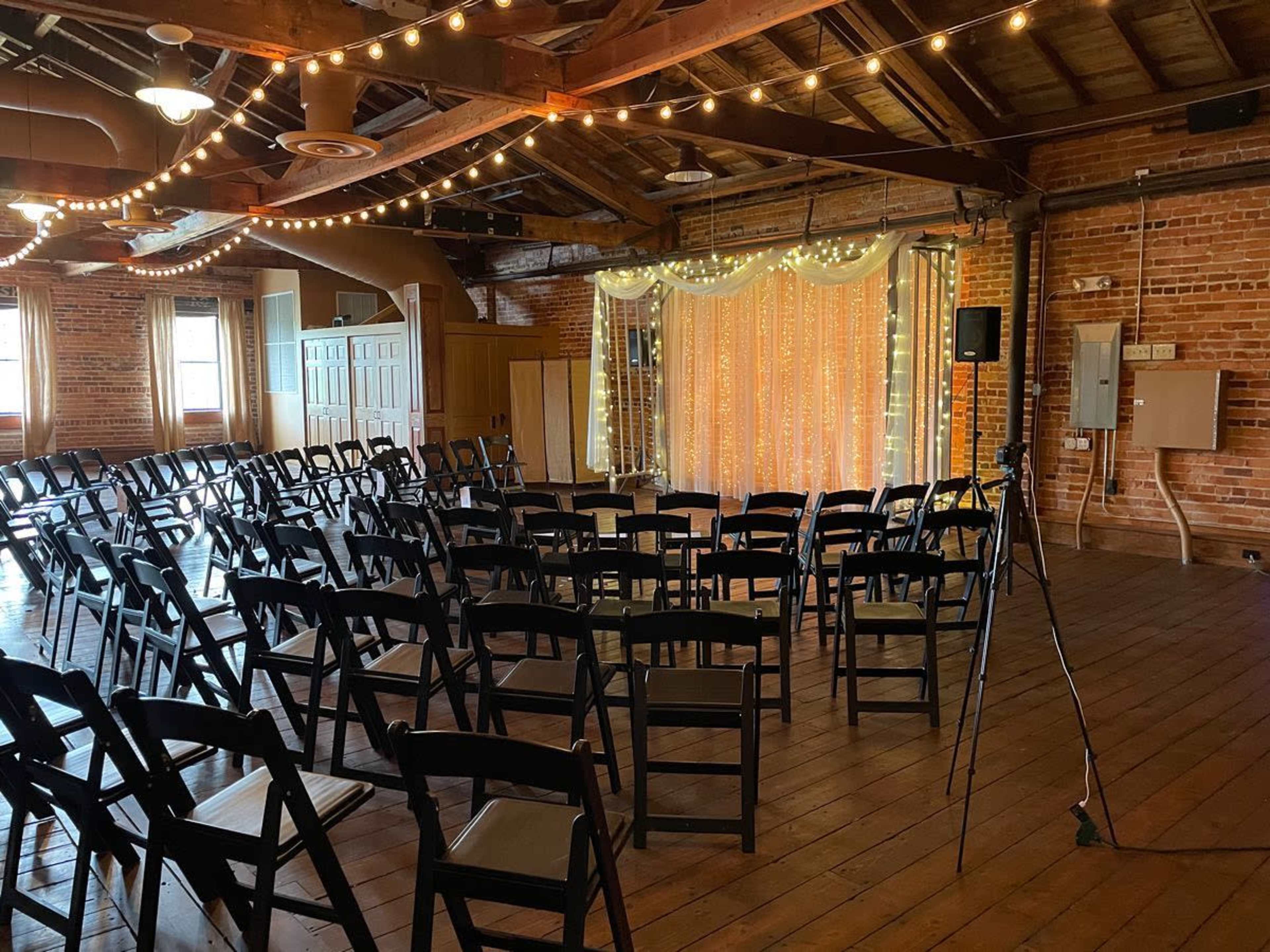 An empty event space with rows of black chairs arranged facing a decorated backdrop and string lights hanging from the wooden ceiling.