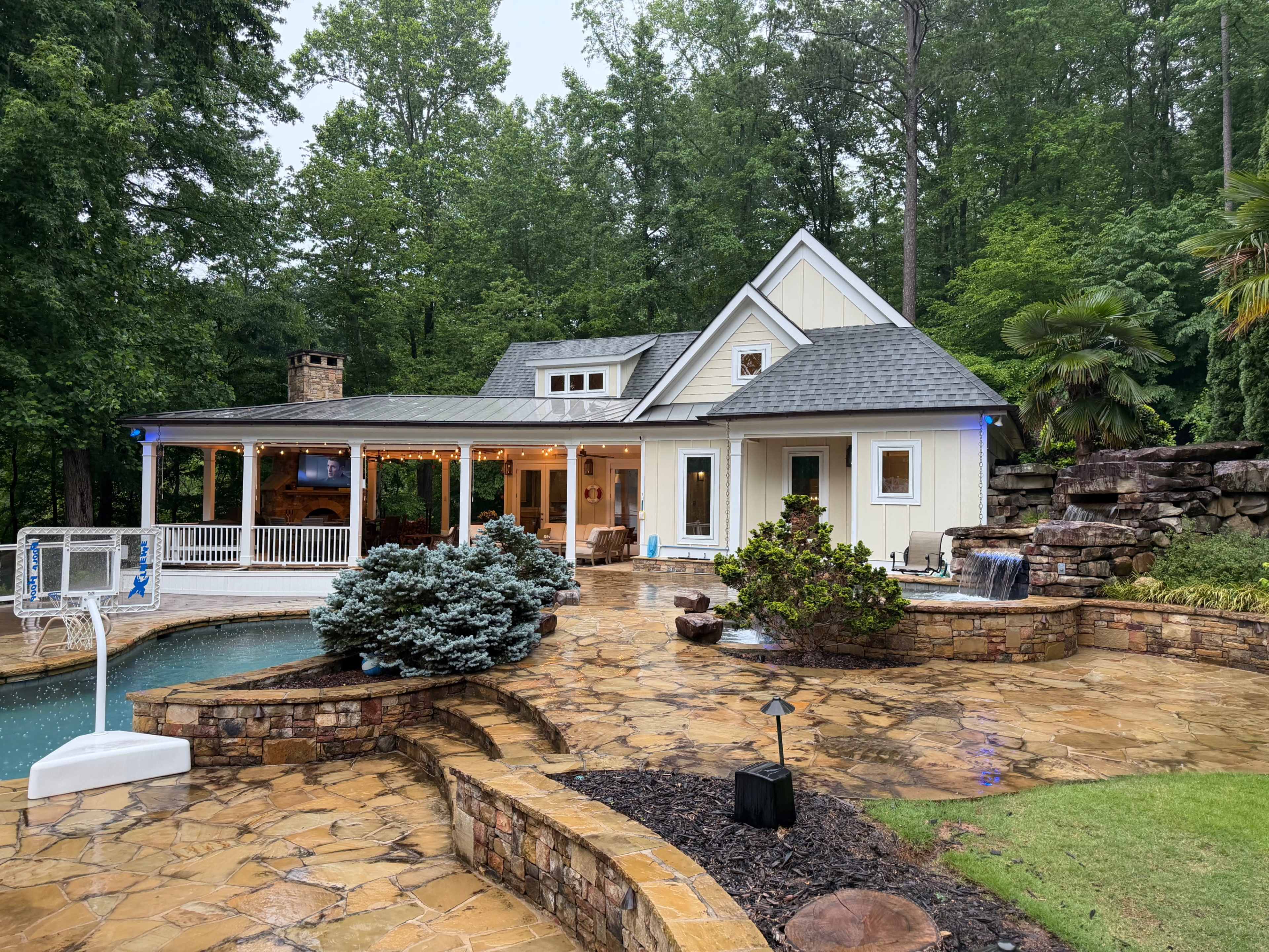 A cozy house with a stone patio, surrounded by trees, features a small swimming pool and a basketball hoop.
