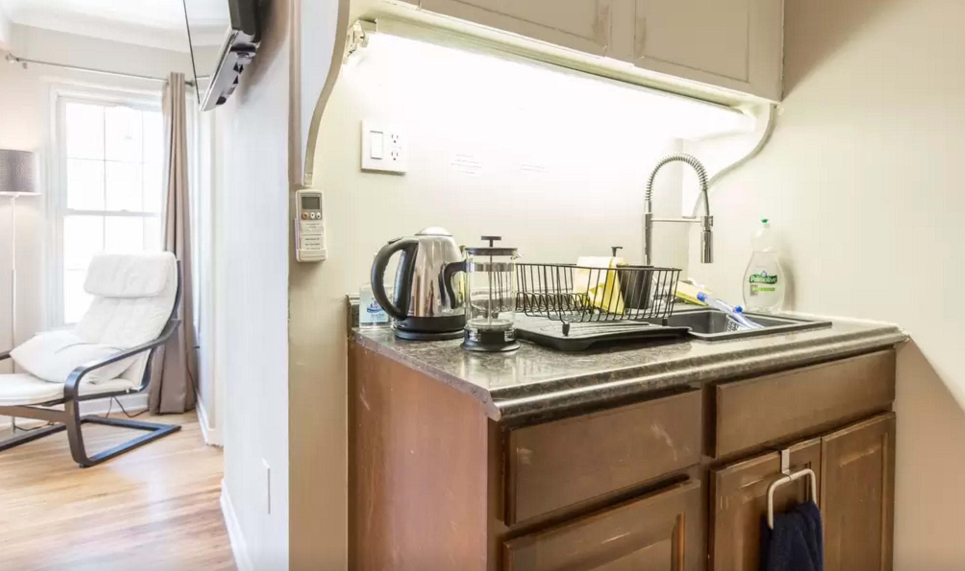 A small kitchen area with a sink, drying rack, kettle, and cleaning supplies, adjacent to a living space with a chair.