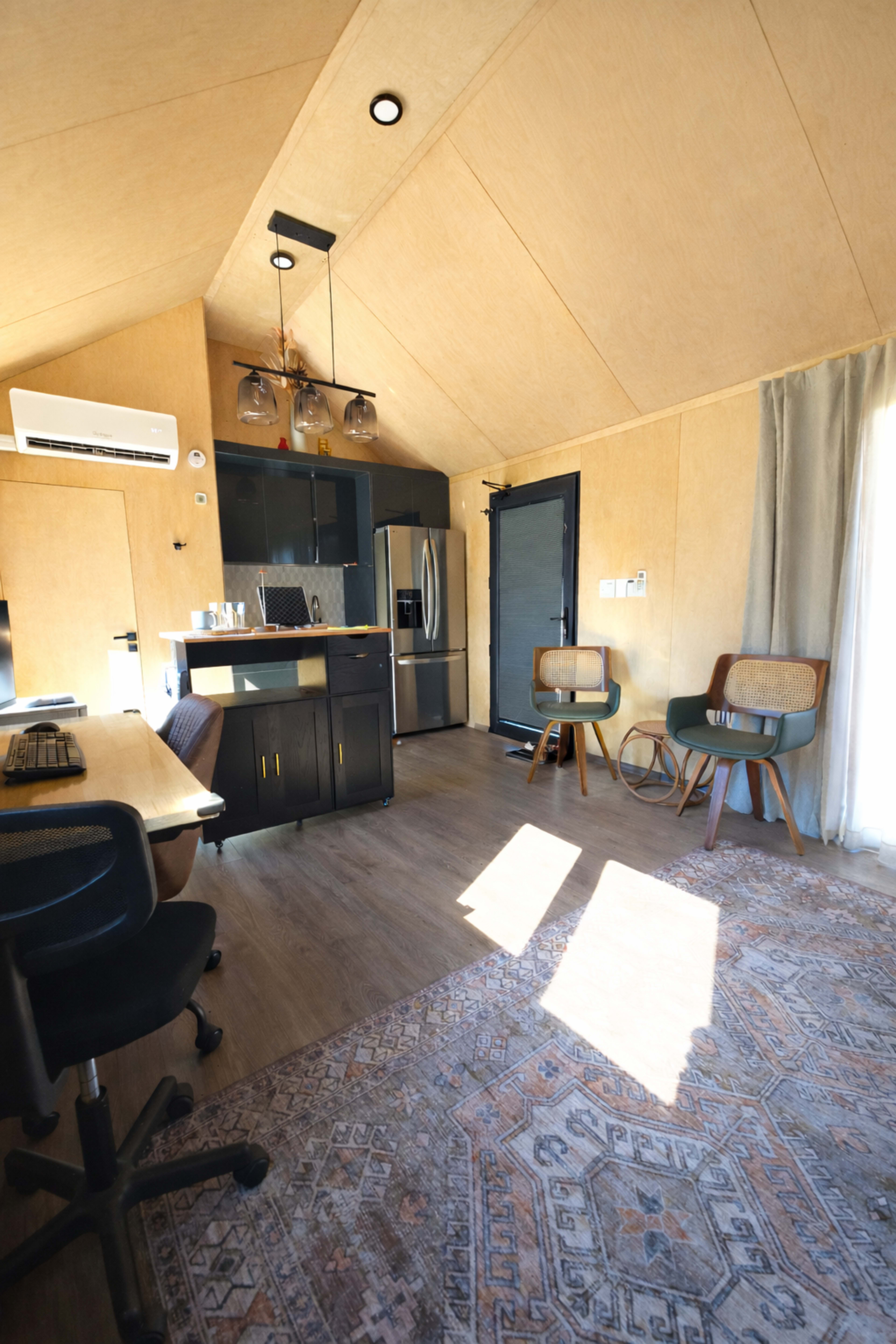 The image shows a compact, modern interior of a furnished tiny house featuring a workspace, kitchen area, and seating.