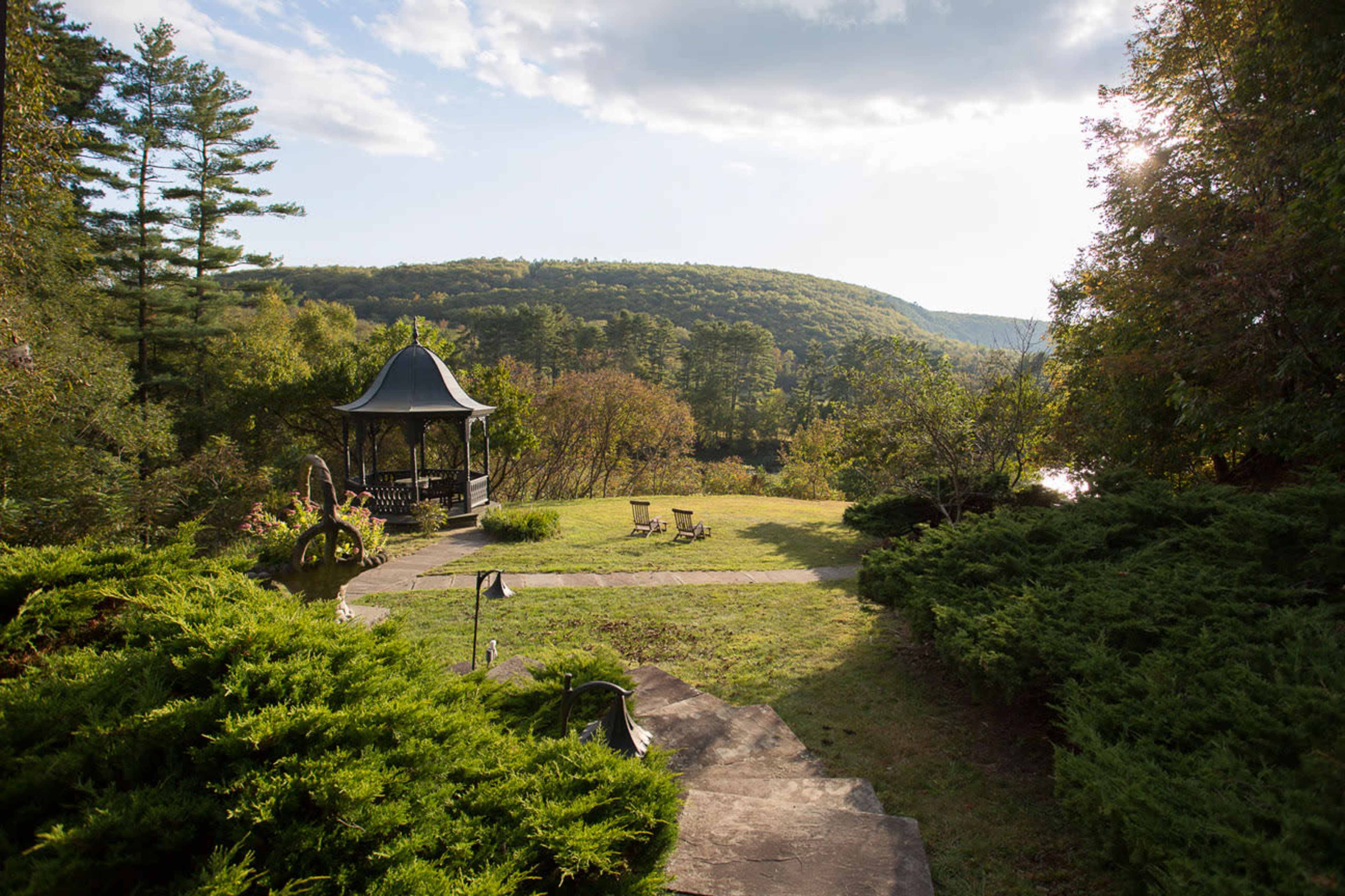 The image depicts a gazebo surrounded by lush greenery, overlooking a vast landscape of rolling hills and a tranquil body of water.