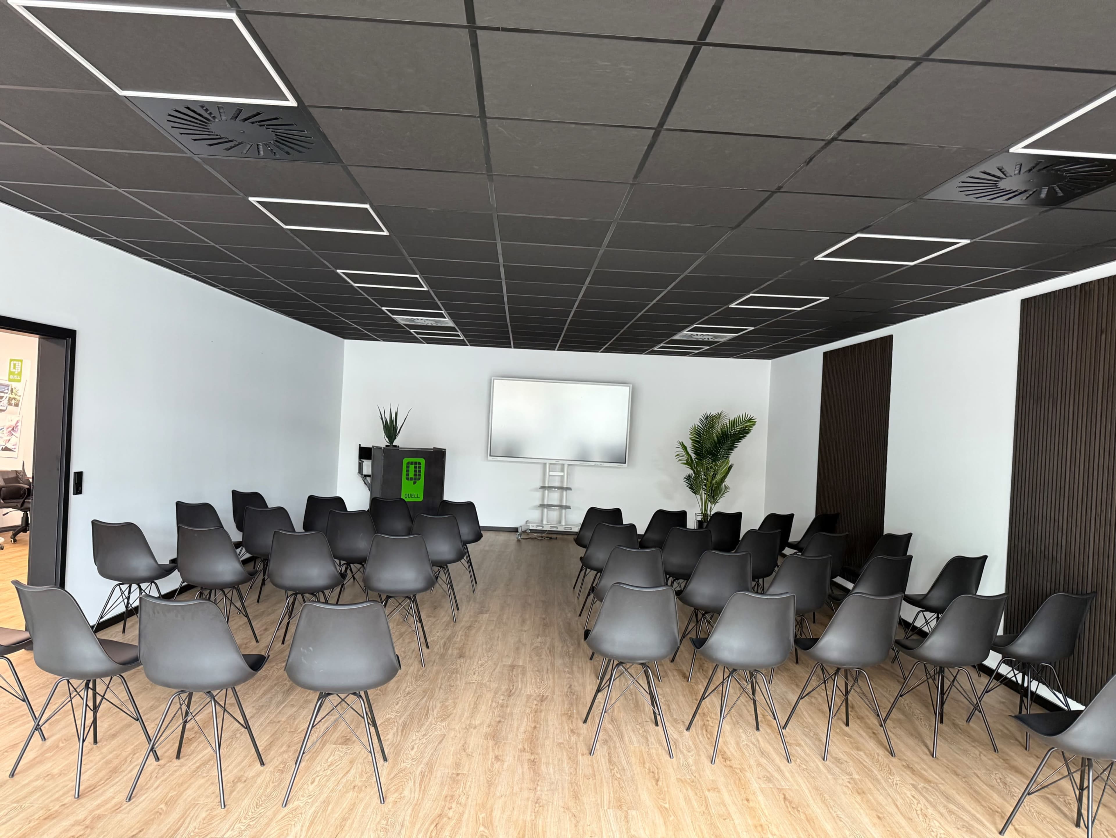 The image shows a conference room set up with multiple black chairs arranged in rows facing a large screen on a white wall.