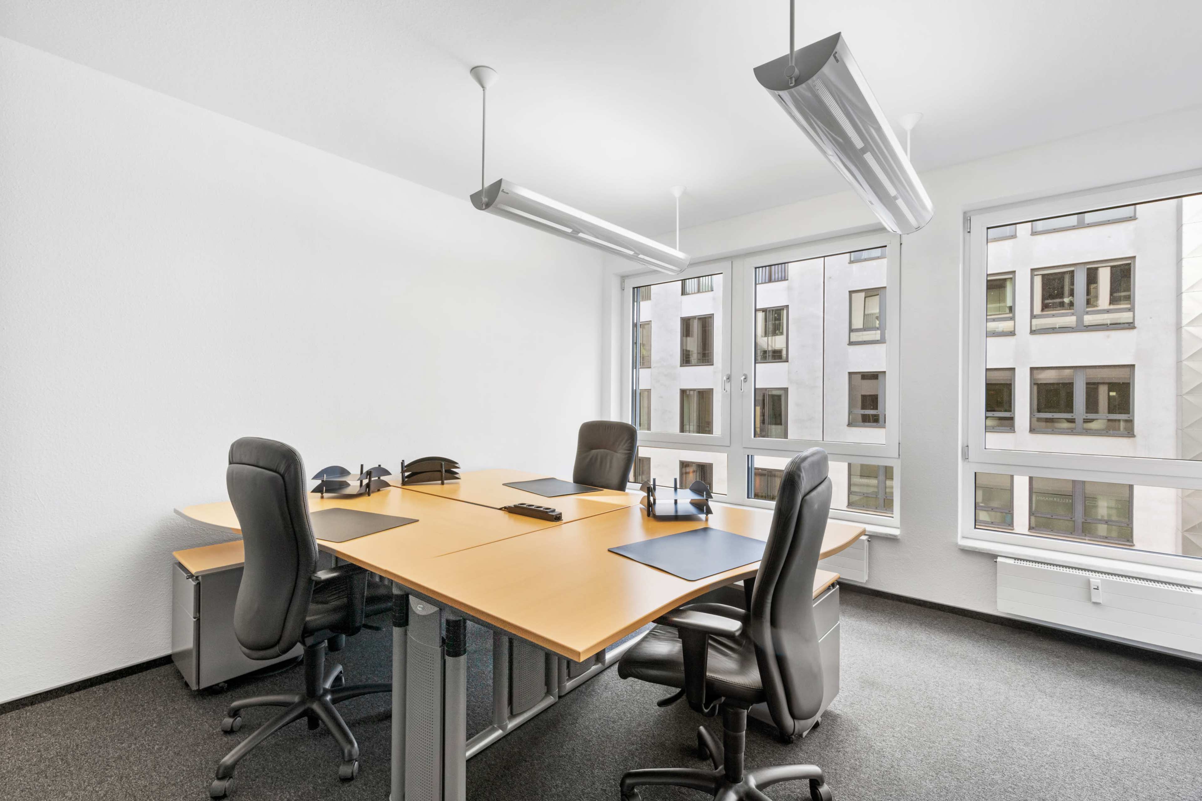 A modern office space features a large wooden table surrounded by black chairs and large windows overlooking a building exterior.
