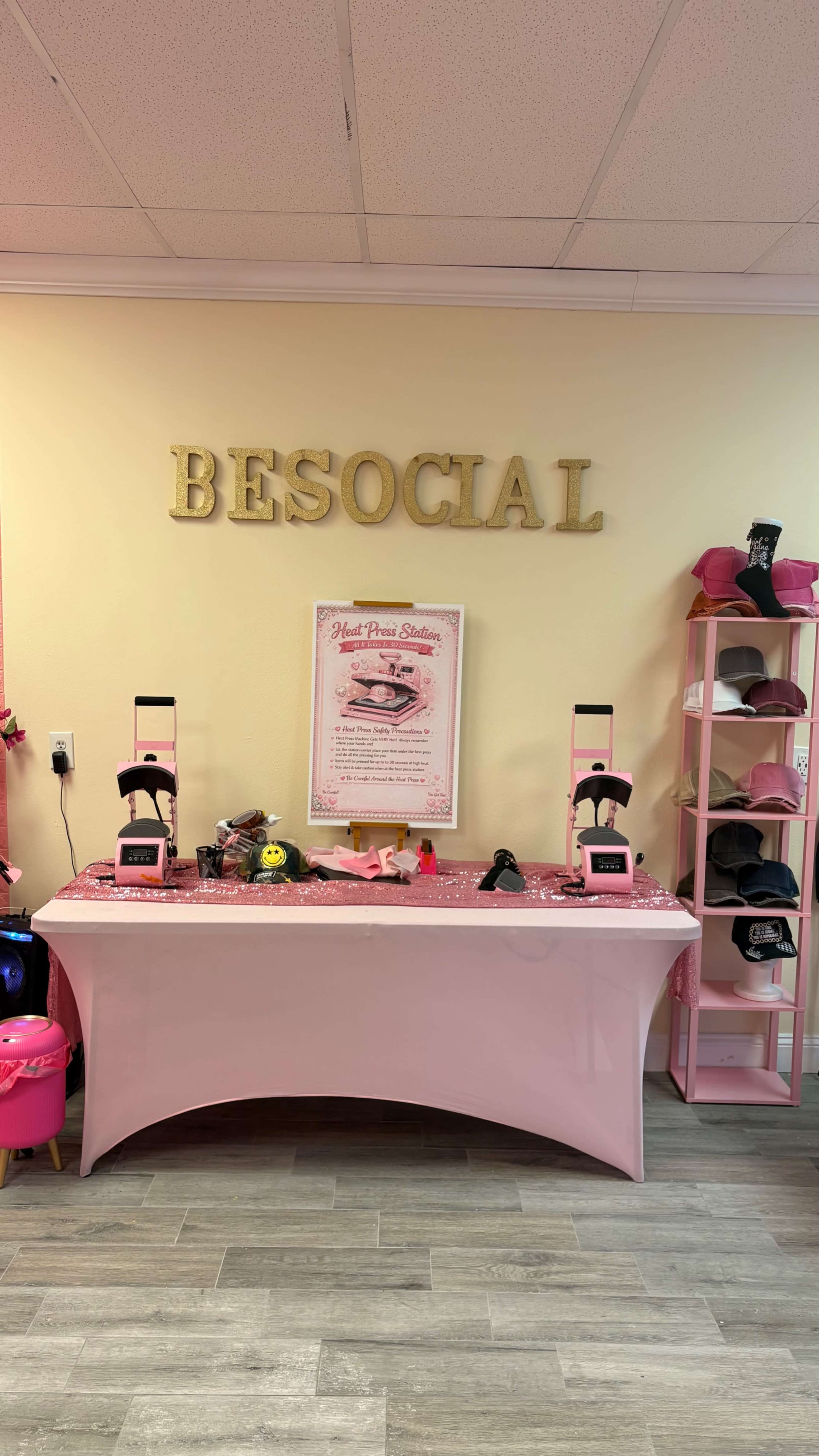 The image shows a brightly colored shop interior featuring a table with various beauty products, a pink display shelf with hats, and the word "BESOCIAL" displayed on the wall.