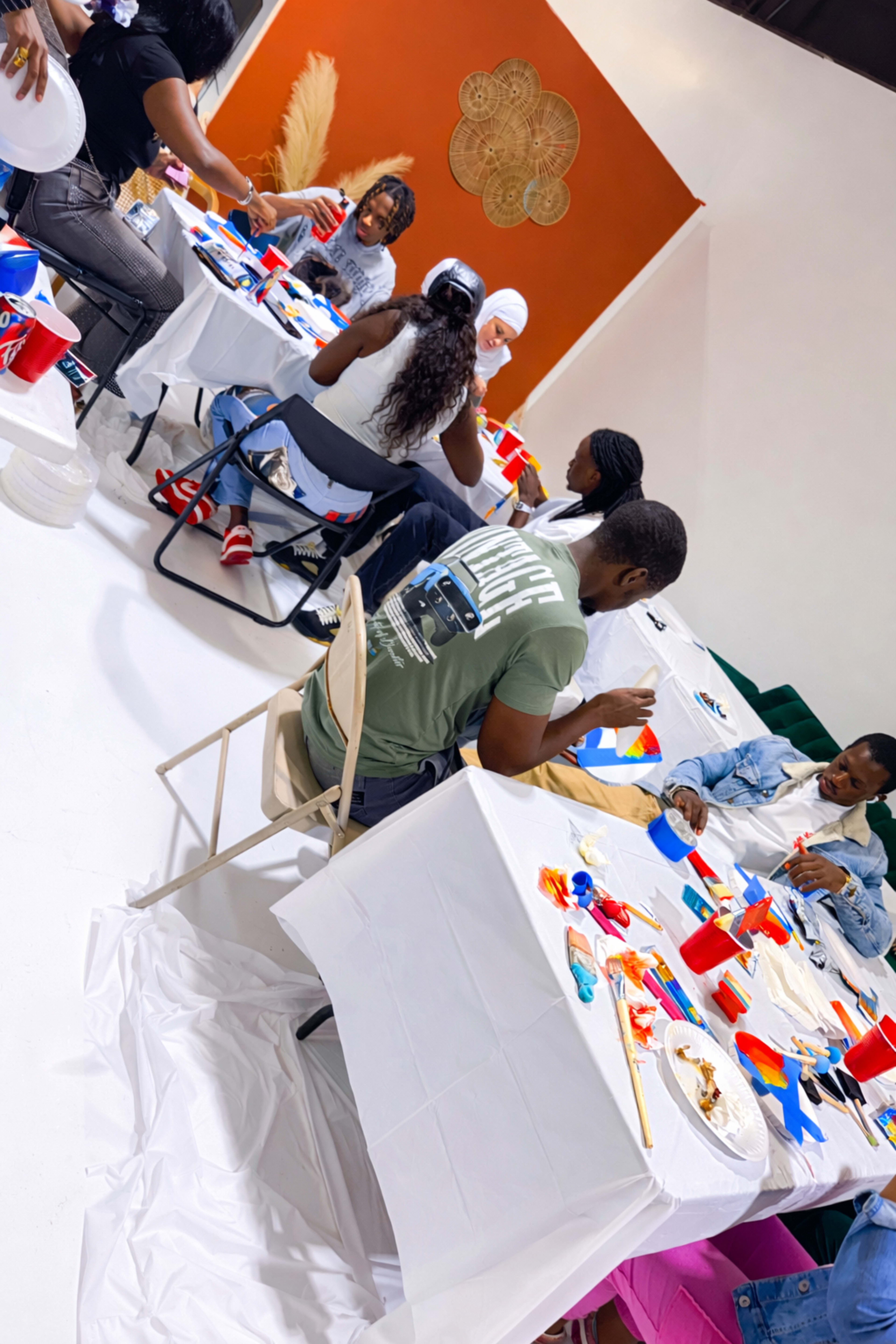 A diverse group of people sits at tables covered with white cloth, engaged in a painting activity within a brightly lit studio.