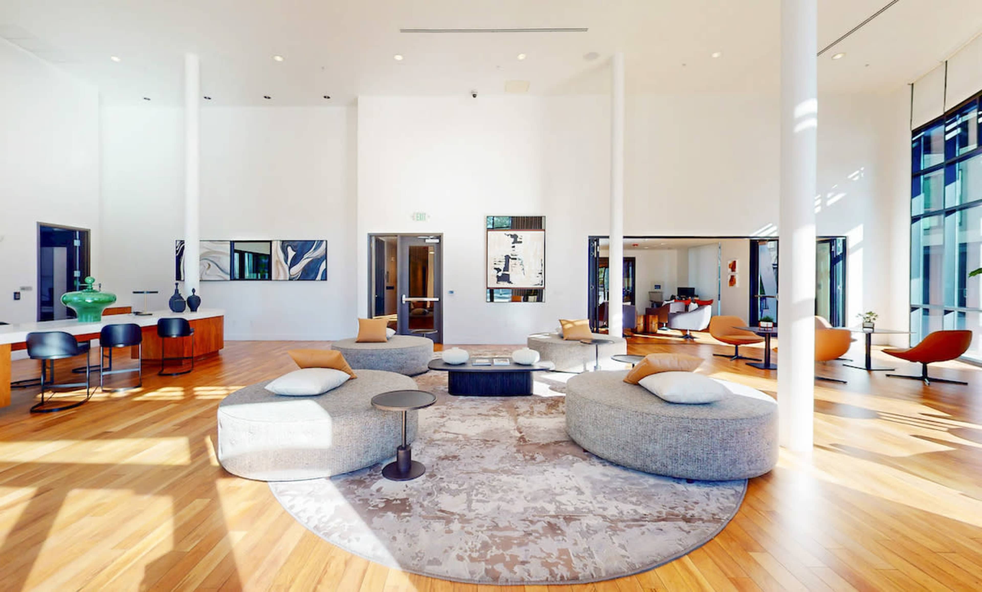 The image shows a modern, open-concept lobby with circular seating areas, wooden floors, and large windows allowing natural light to enter.