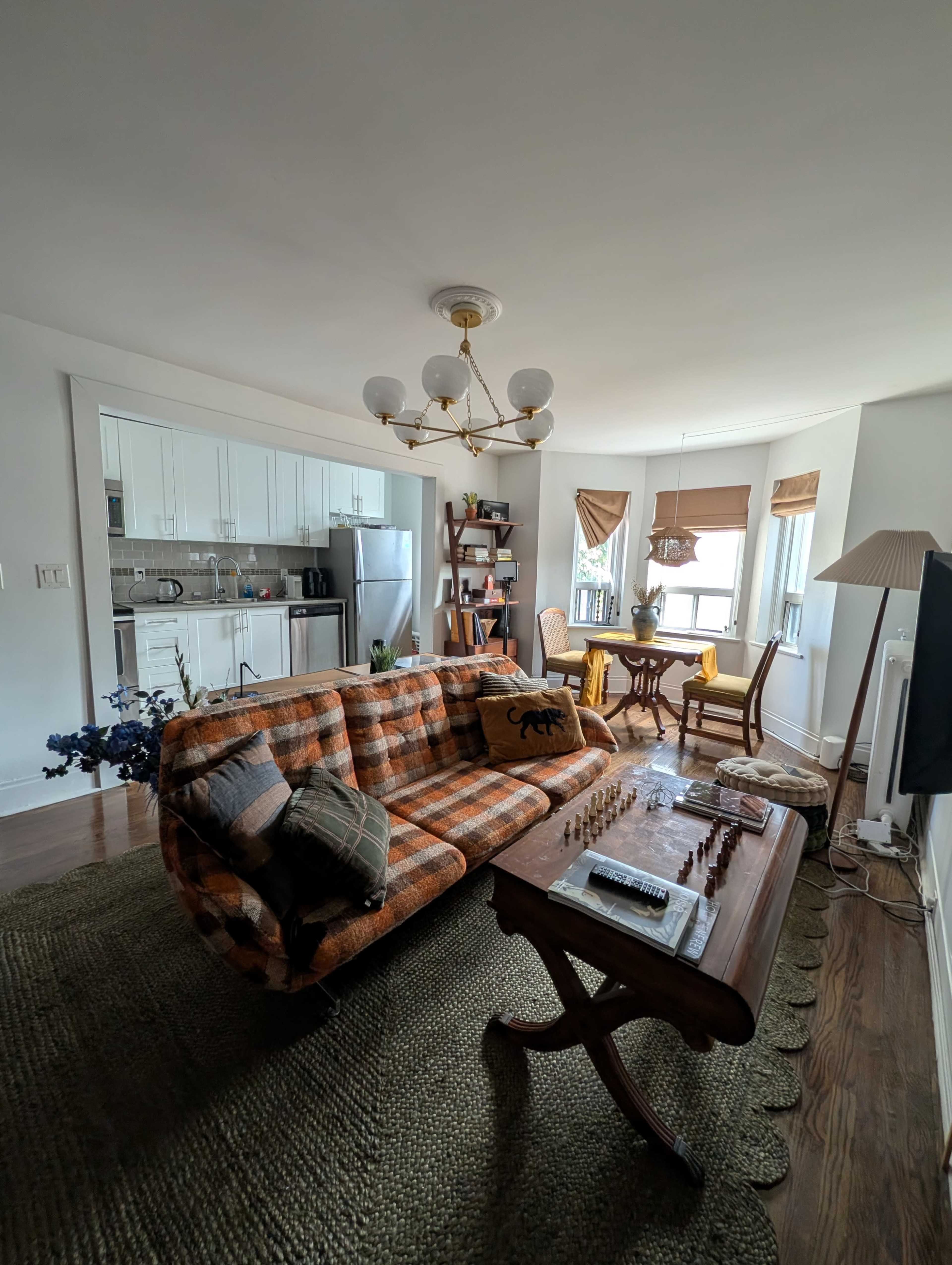 The image shows a cozy living room featuring a plaid sofa, a wooden coffee table with a chessboard, and a dining area with a round table and chairs, all set in a well-lit space.