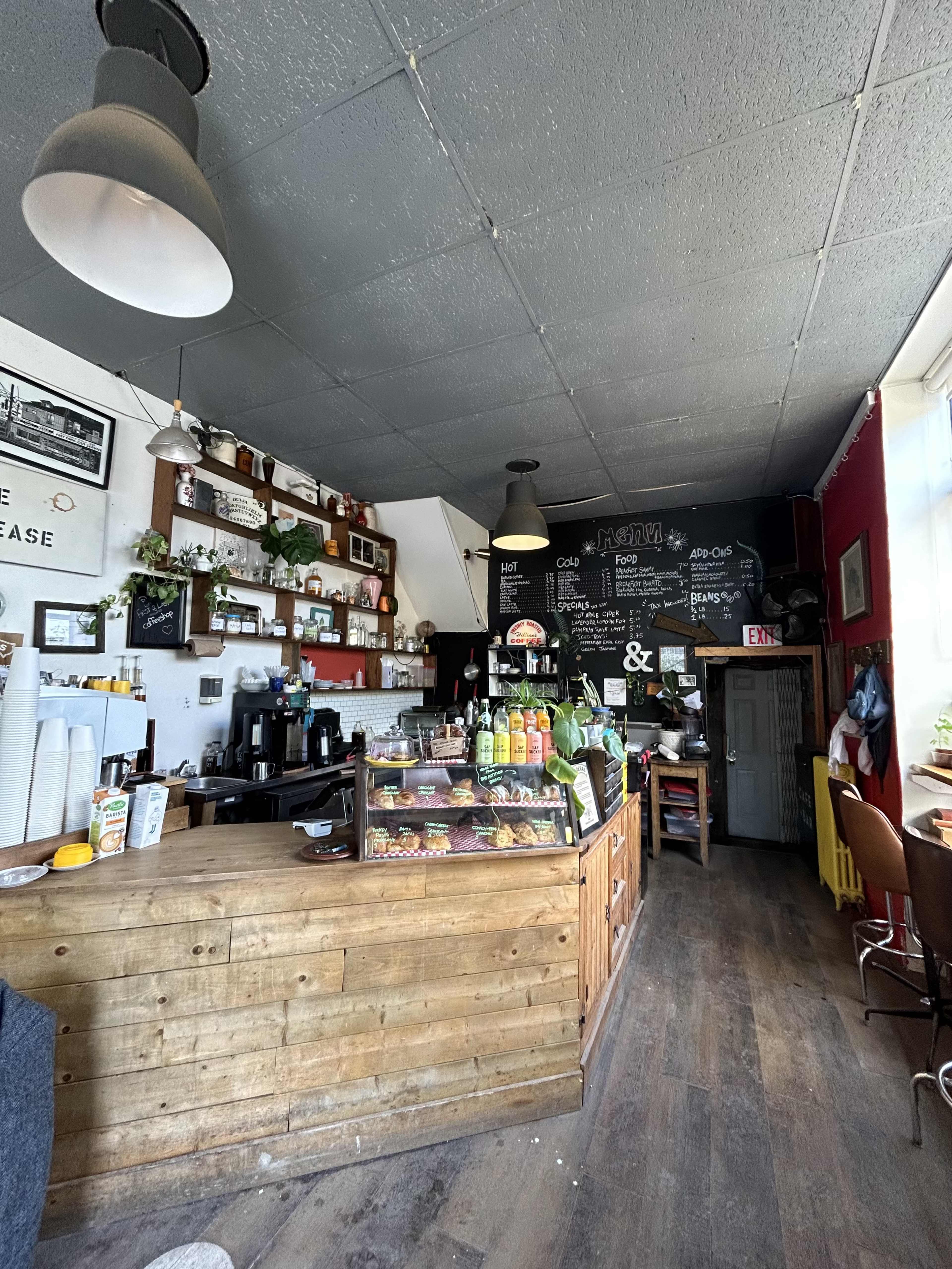 A cozy café interior featuring a wooden counter with various drinks and pastries, surrounded by shelves adorned with plants and decorative items.