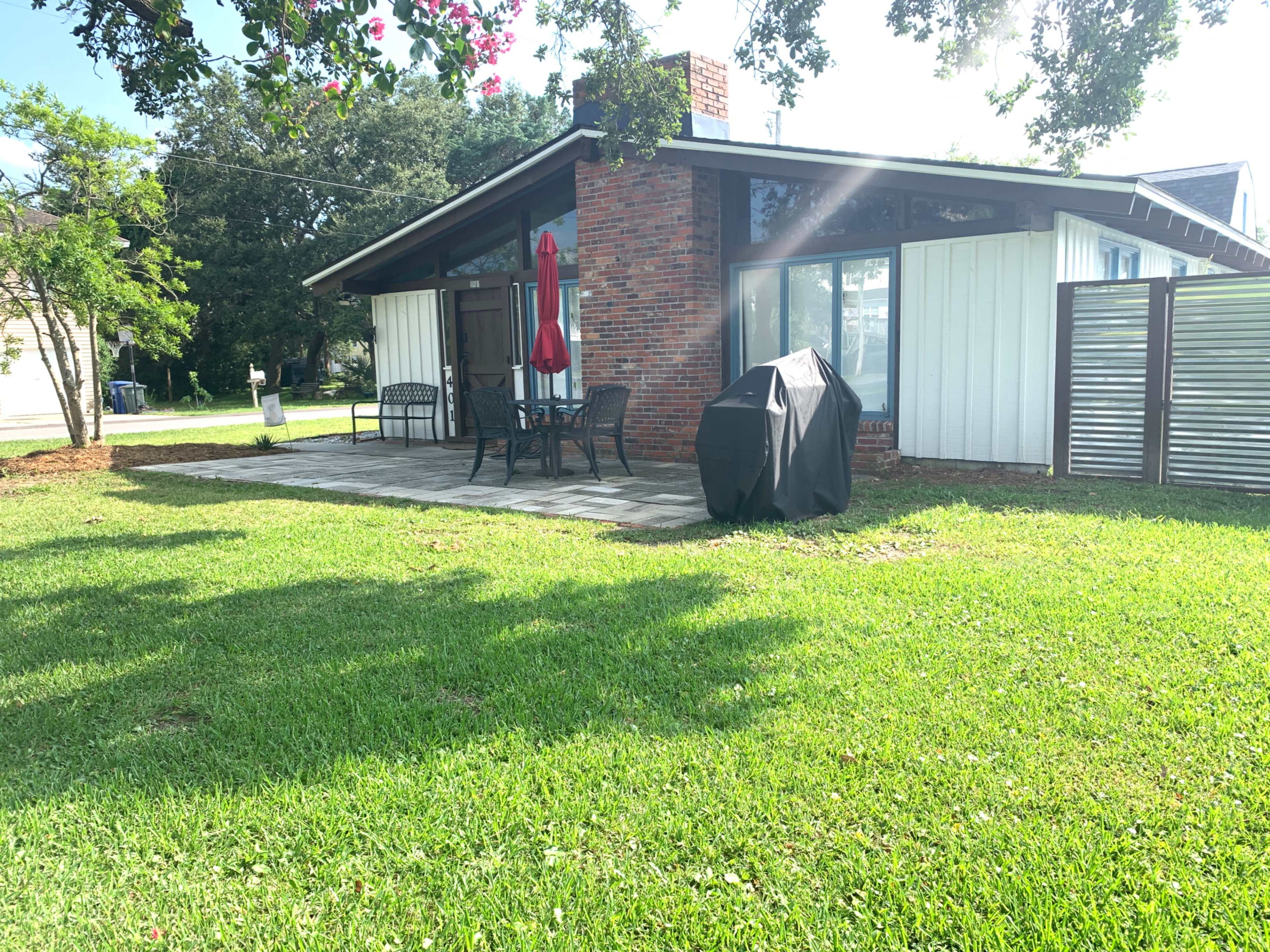 The image shows a backyard patio with a set of chairs and a table under a red umbrella, next to a black grill, and surrounded by green grass.