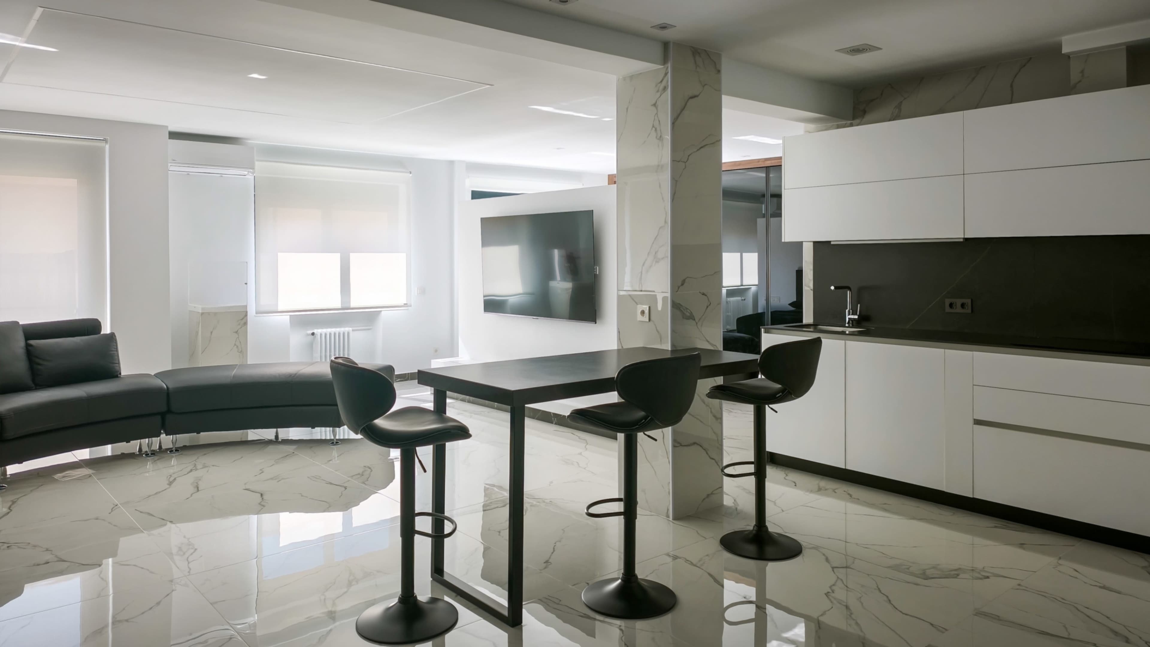 The image shows a modern kitchen and living area with a marble floor, a bar table with three stools, and sleek white cabinetry.