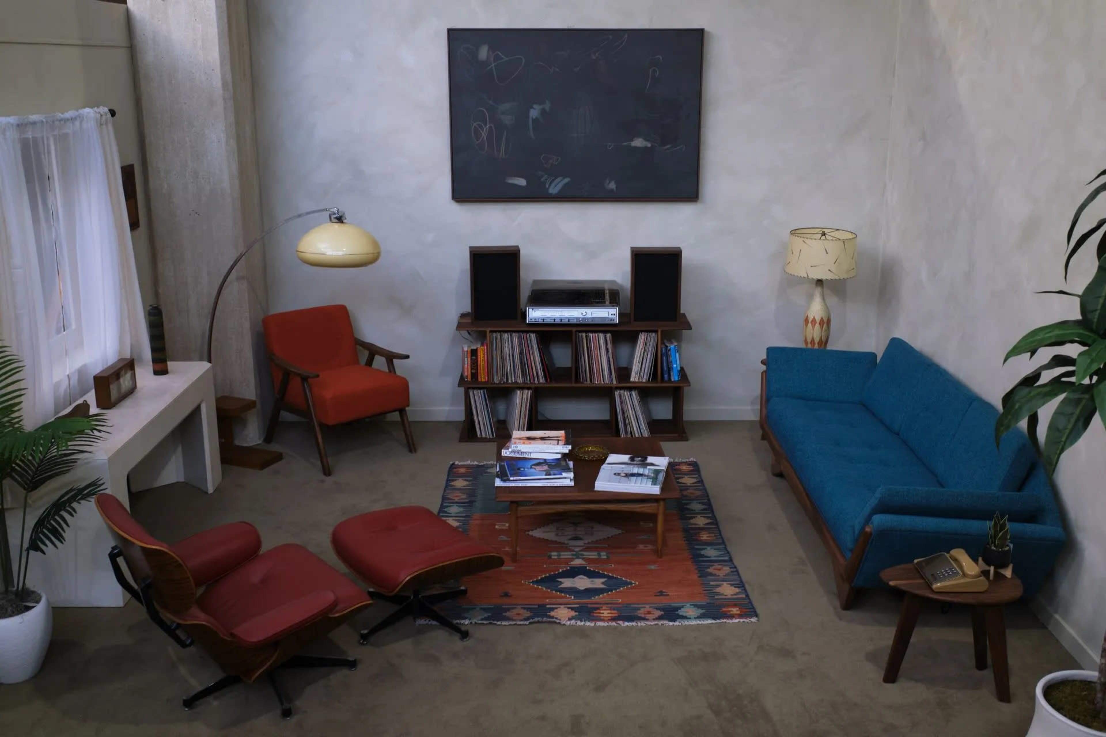 The image depicts a mid-century modern living room featuring a blue sofa, red chairs, a wooden coffee table, and a record player on a shelf against light-colored walls.