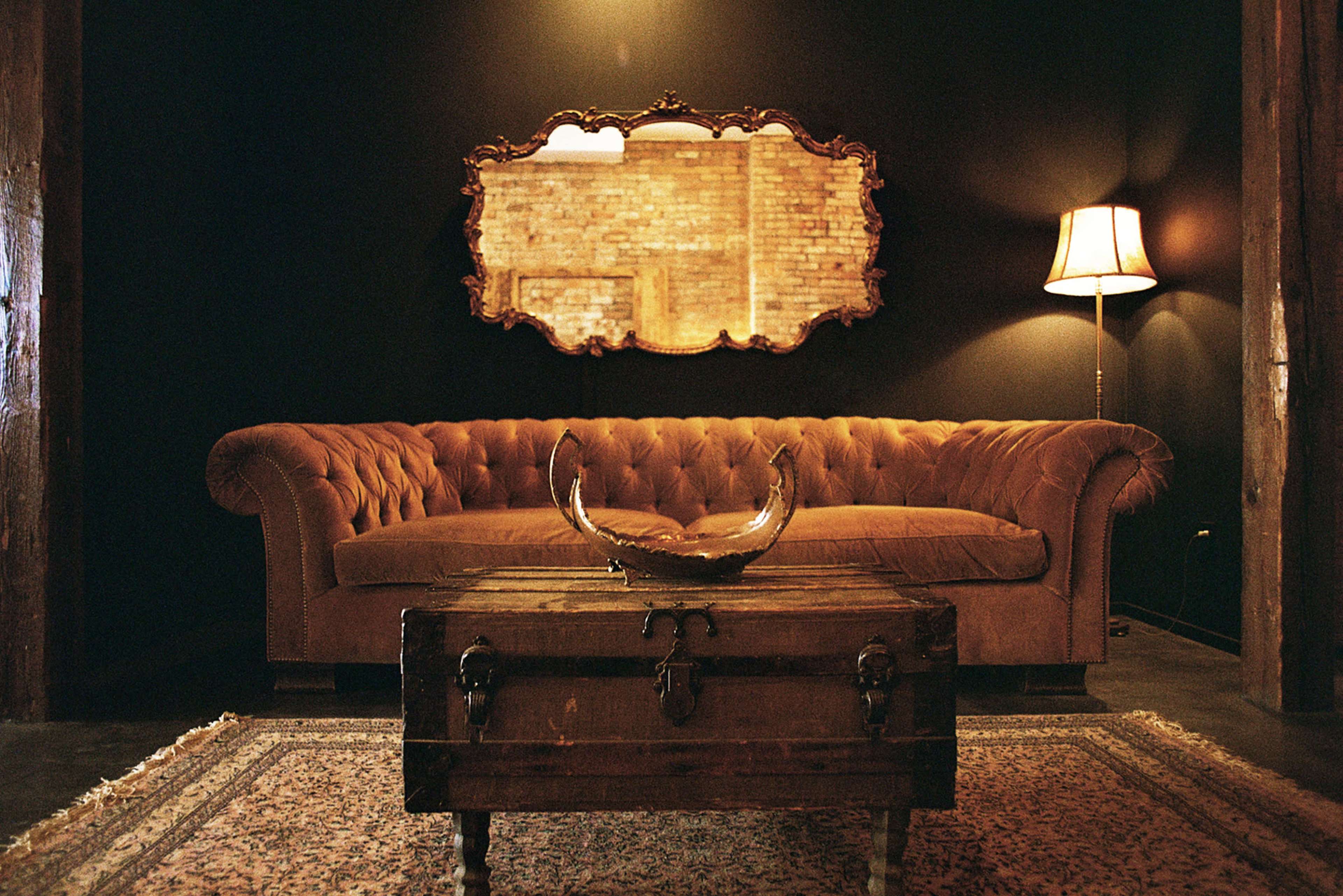 The image shows a vintage living room featuring a tufted brown couch, a wooden coffee table with decorative items, a large mirror reflecting a brick wall, and a standing lamp.