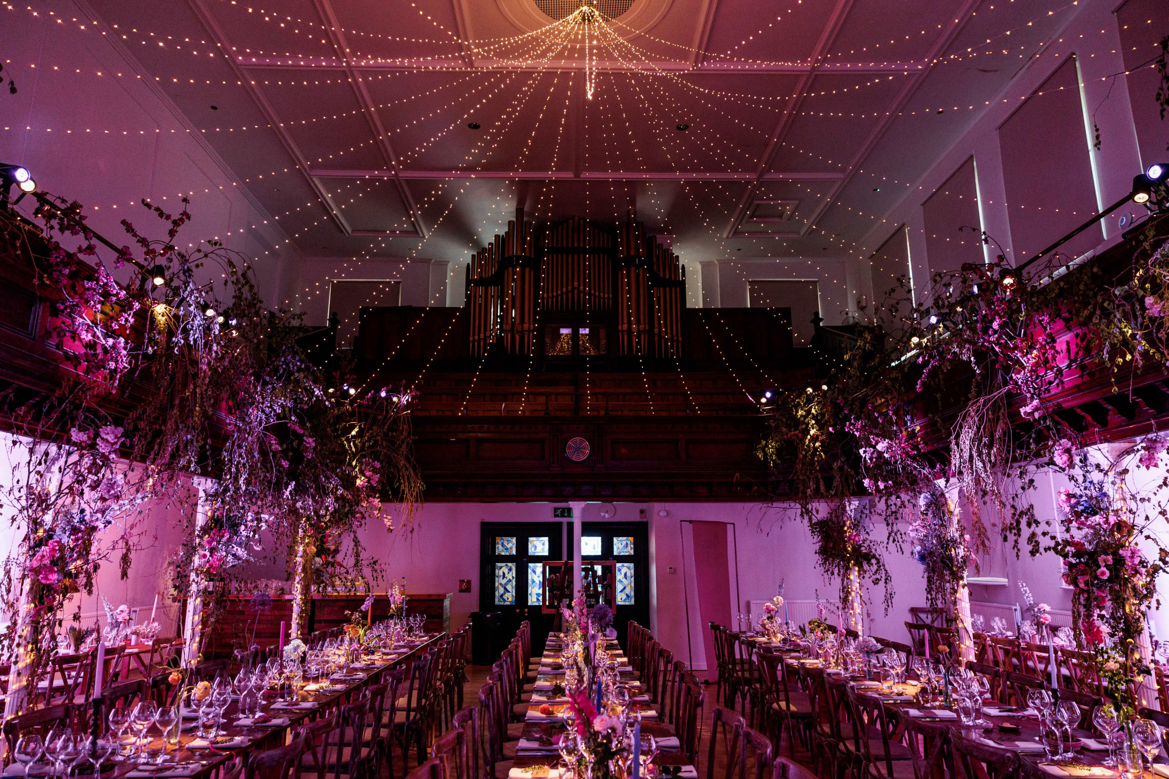 A banquet hall is adorned with floral arrangements and string lights, featuring long dining tables set for a formal event.