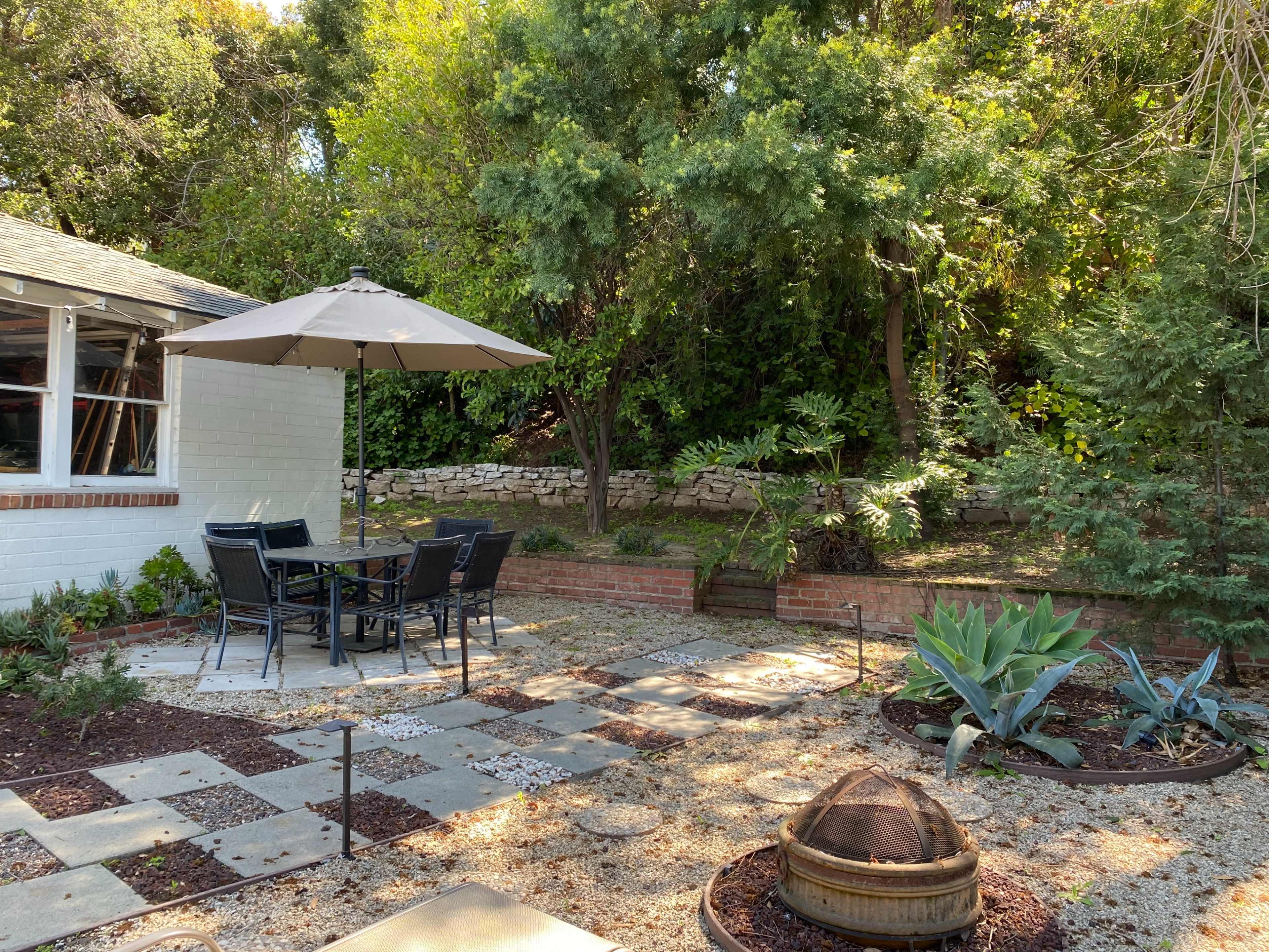 A patio area features a table with chairs beneath an umbrella, surrounded by stone pathways, planters, and various plants.