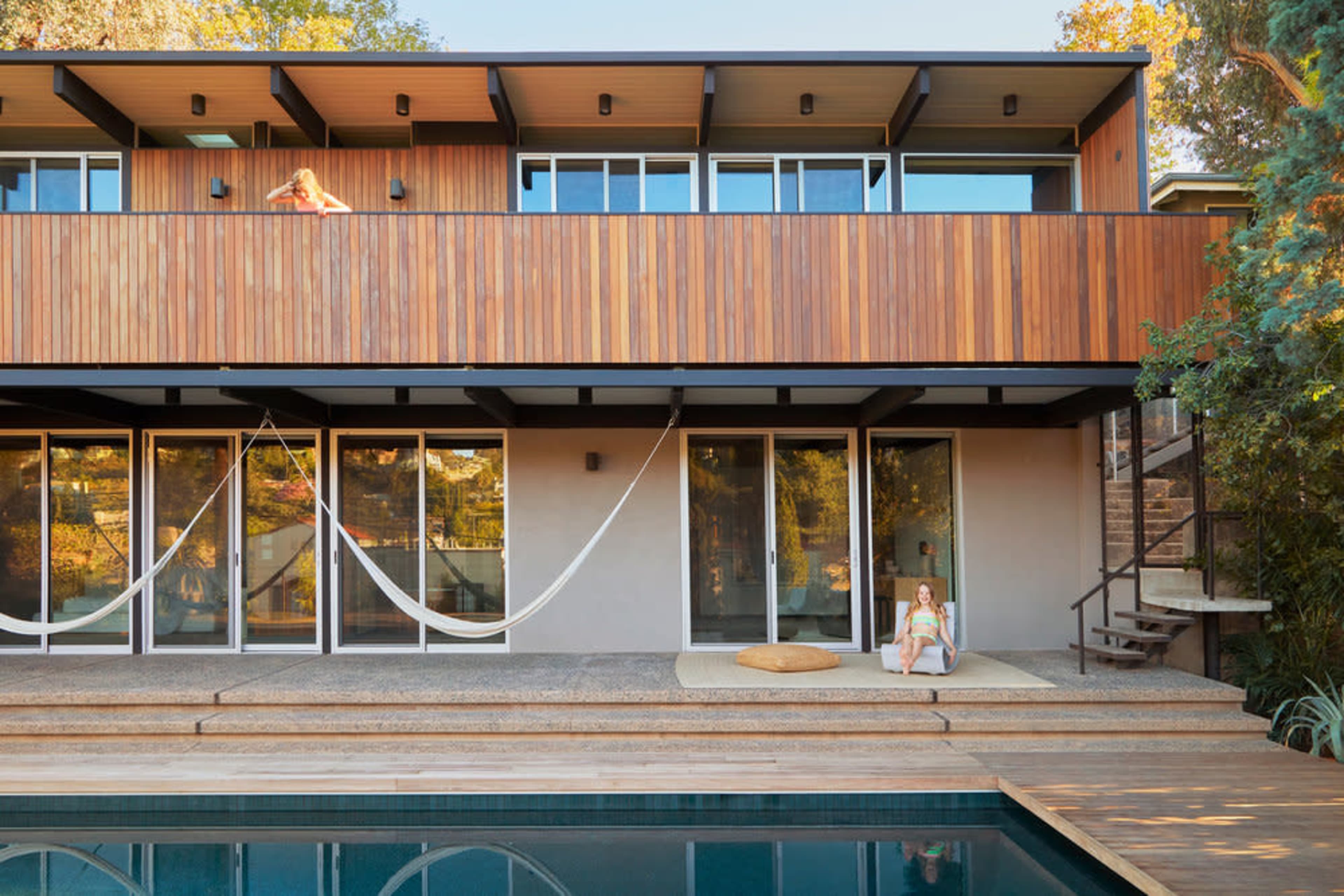 A modern home features a wooden balcony overlooking a swimming pool, with two people relaxing in the outdoor space.