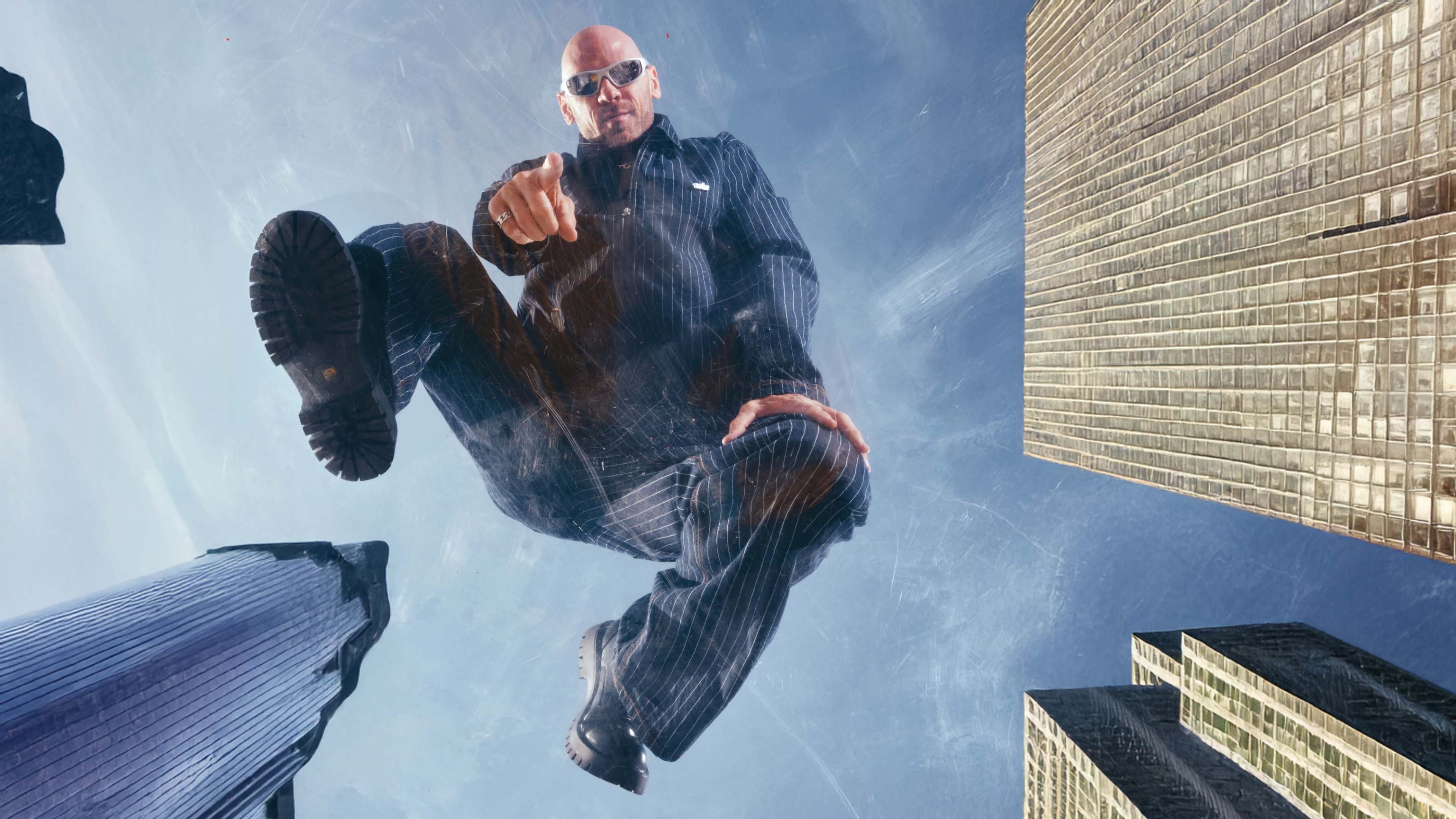 A man in sunglasses is jumping over a reflective surface with tall skyscrapers visible around him.