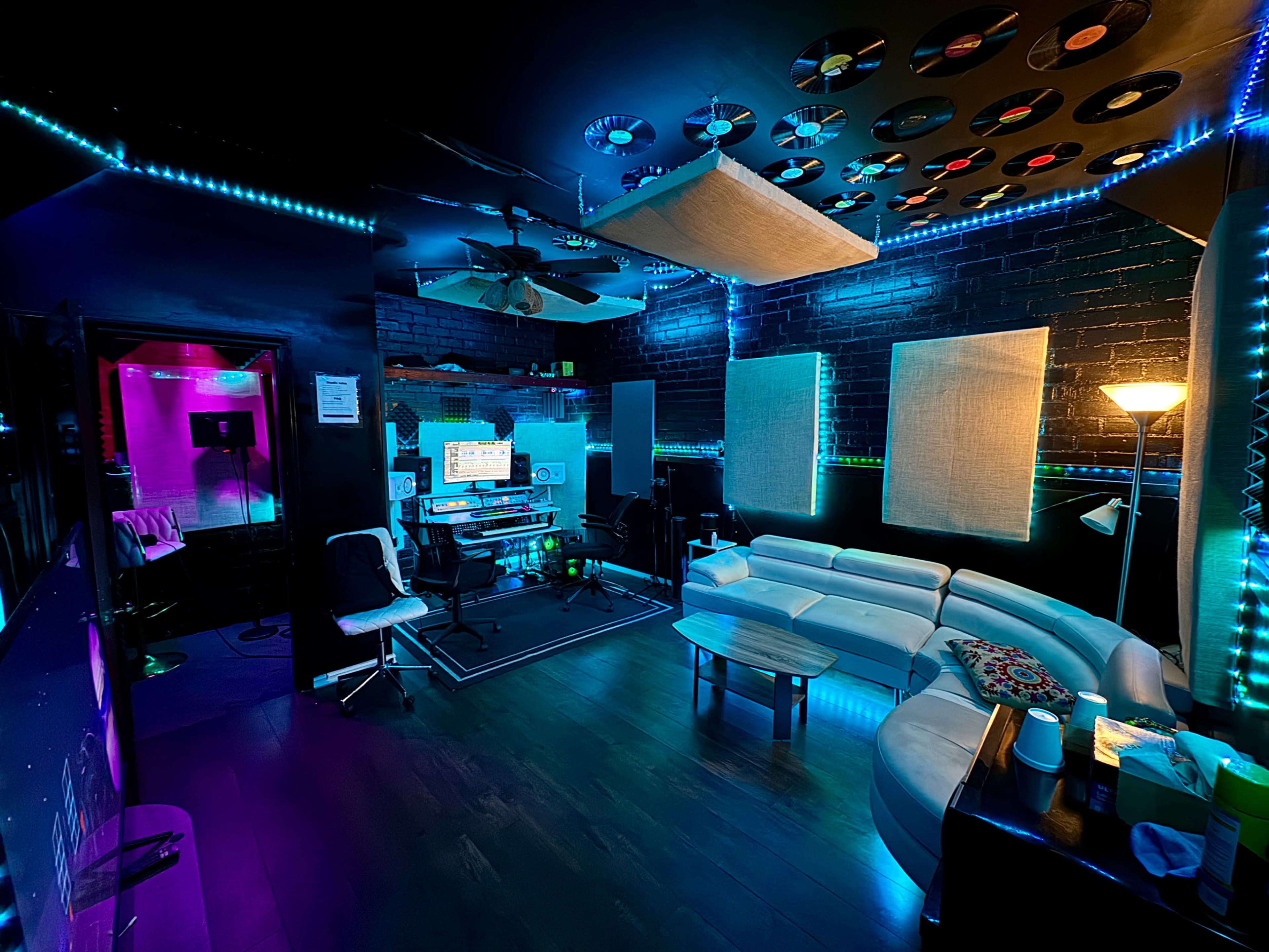 The image shows a music recording studio featuring colorful LED lights, soundproofing panels, a seating area with a white couch, and various music equipment.