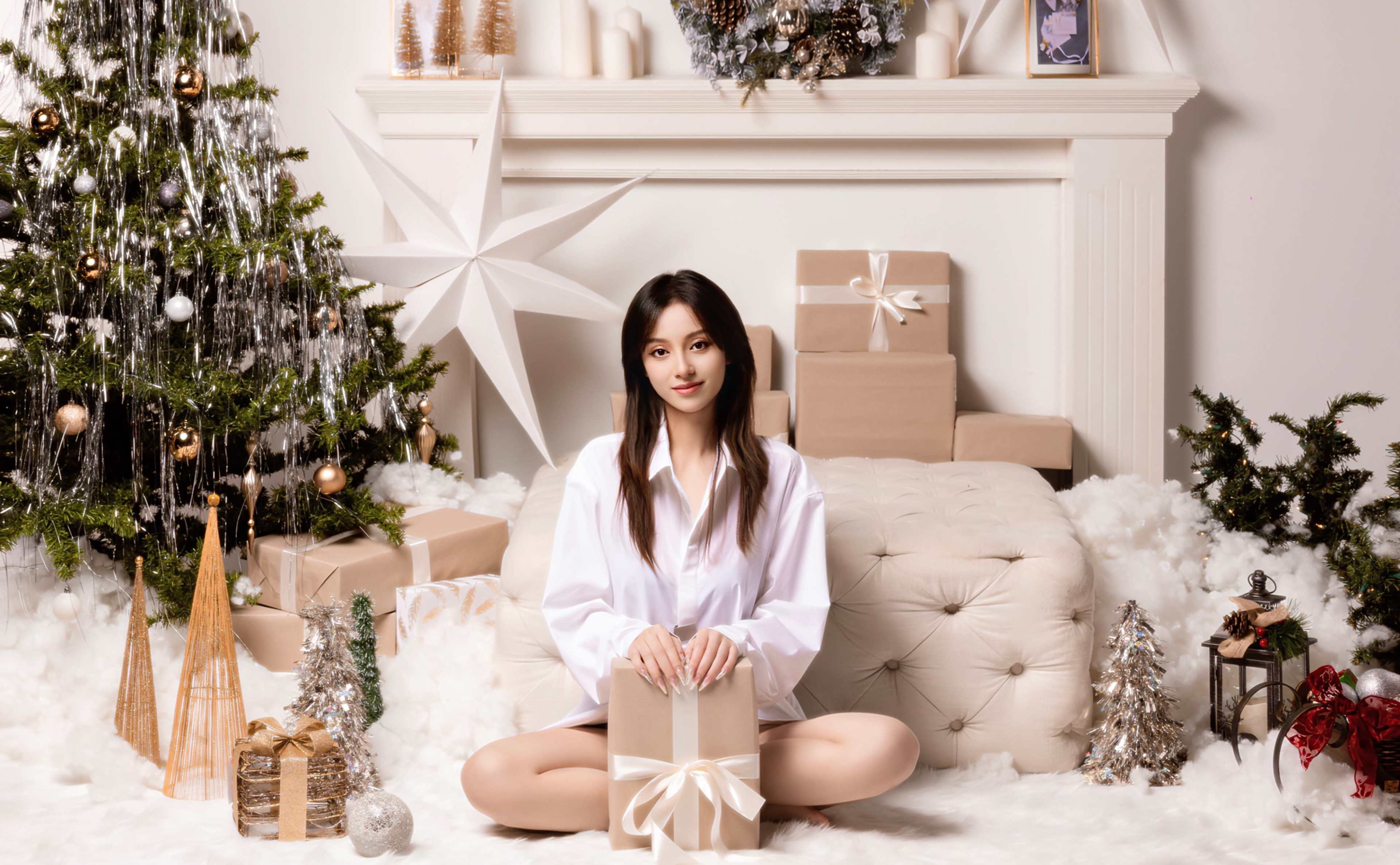 A woman in a white shirt sits on a fluffy rug surrounded by holiday decorations, including a Christmas tree, presents, and silver-colored ornaments.