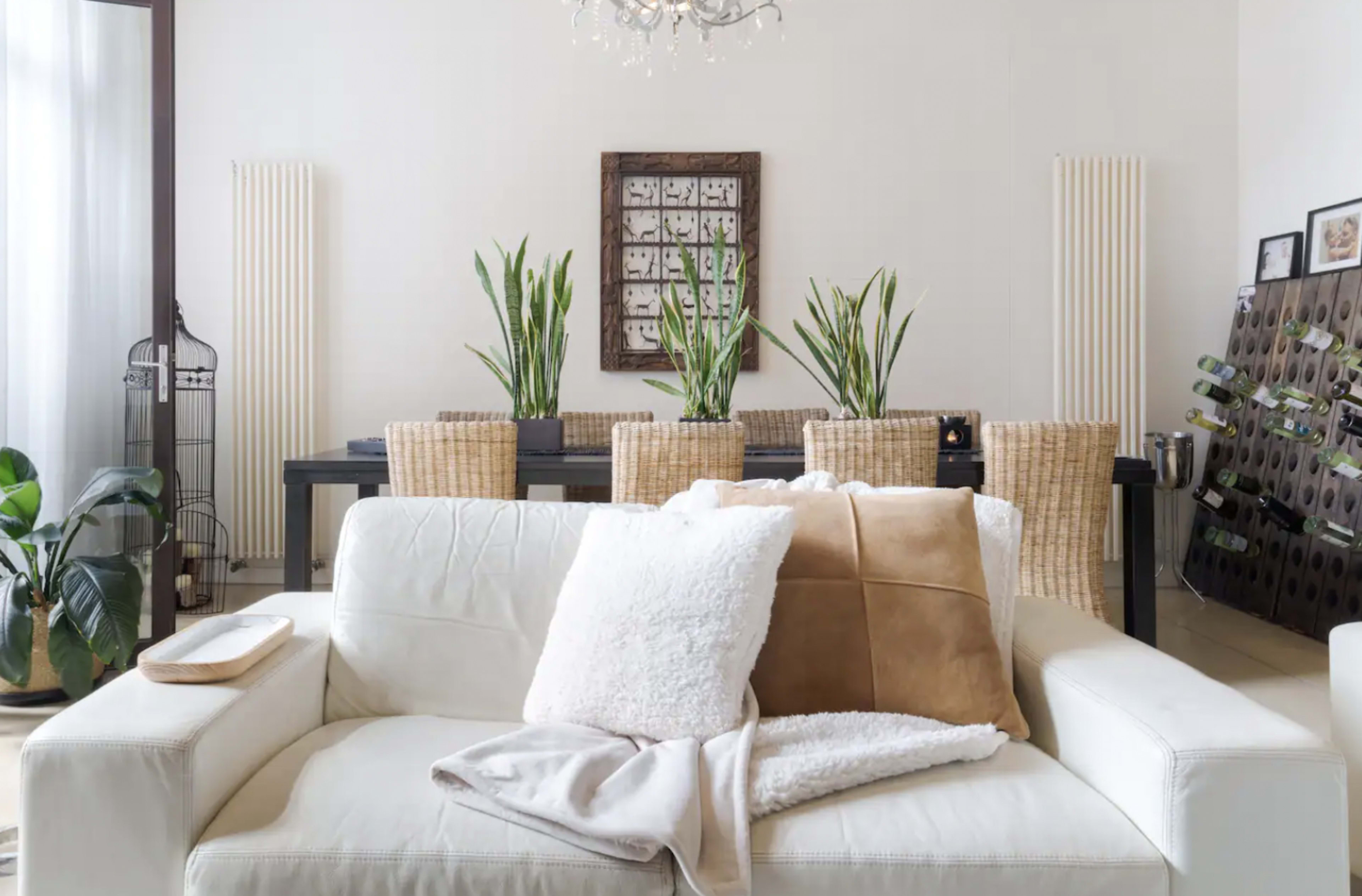 A modern living room features a white sofa with decorative pillows facing a dining area with a table surrounded by woven chairs and plants.