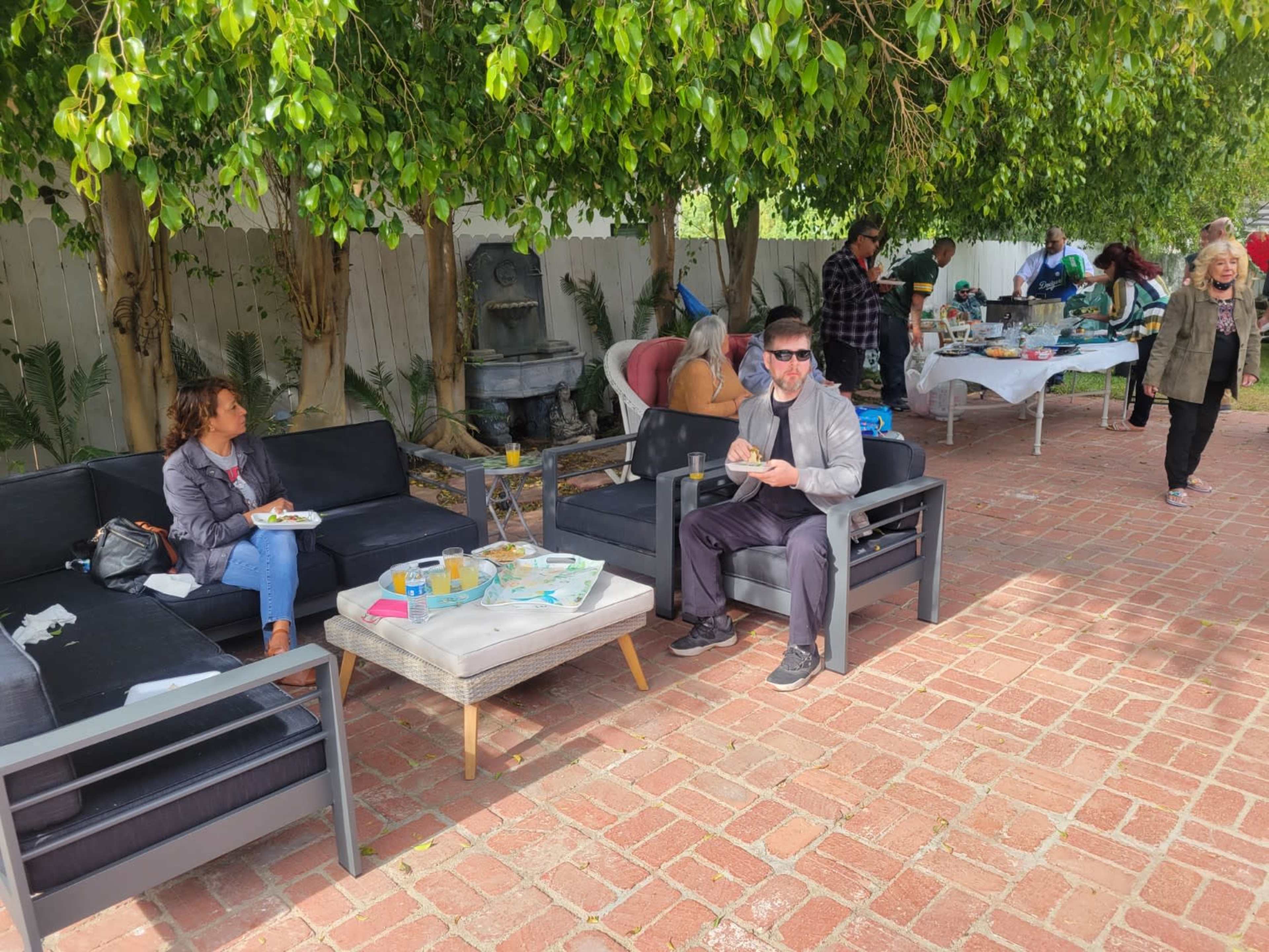 A group of people socializes in a garden setting with outdoor seating, enjoying food and drinks under greenery.